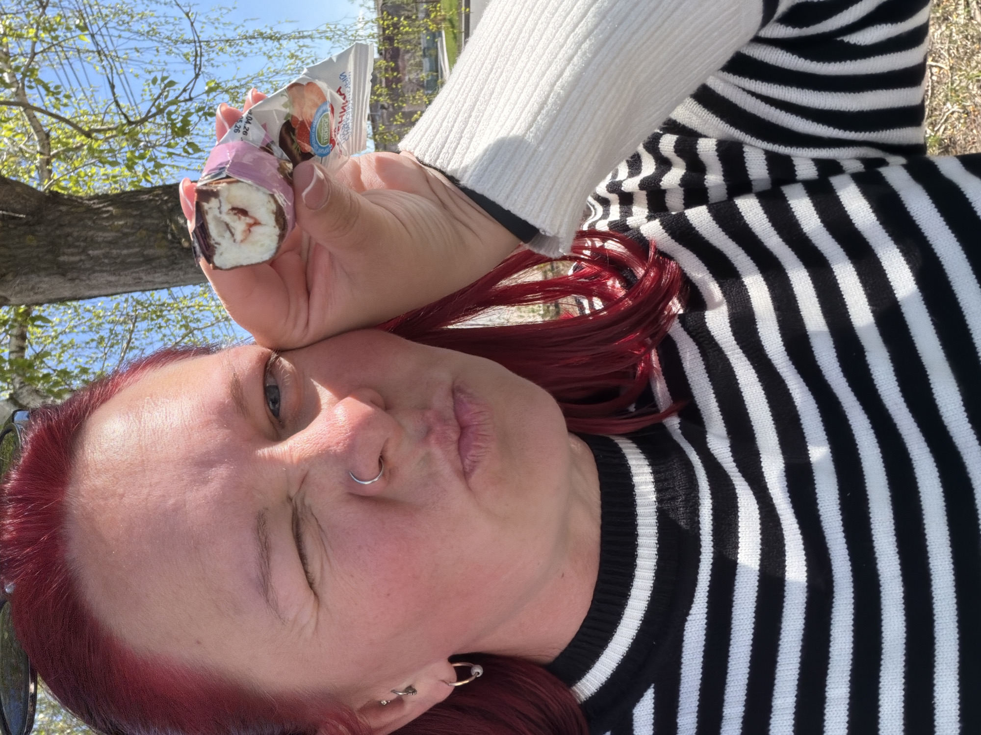 The image depicts a woman with red hair outdoors on a sunny day. She is wearing a black and white striped sweater with a white undershirt. The woman is holding a chocolate-covered cream cheese bar, possibly a "syrki" or "glazirovanny syrok," a popular Russian dairy snack. The background includes a glimpse of trees with budding green leaves, suggesting springtime. The presence of buildings with visible architectural details may indicate the location is within a city.
The image was taken in Chaikovsky, Russia.
