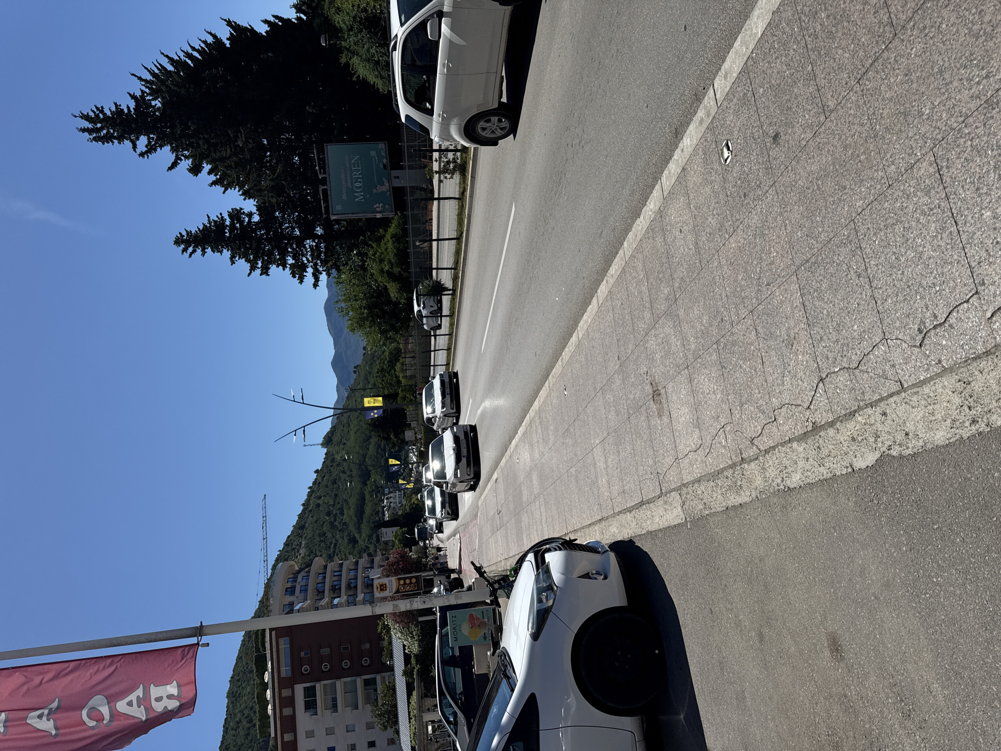 This is a medium-angle, vertical outdoor shot of a street scene in Budva, Montenegro. In the foreground, a white Toyota Yaris is parked on the left side of the image, its front bumper and wheel visible. To its right, a sidewalk paved with textured granite tiles runs alongside the road. The road itself features white lane markings and is populated by several vehicles, including a white SUV on the right side, a white sedan, and other cars further down the street. The background displays a lush green hillside dotted with trees and buildings. A tall, thin flagpole with a maroon banner hangs on the far left. A billboard with the text "MOGREN" is visible in the distance above a row of trees. The sky is a clear, bright blue. The overall scene depicts a sunny day in a coastal city.
