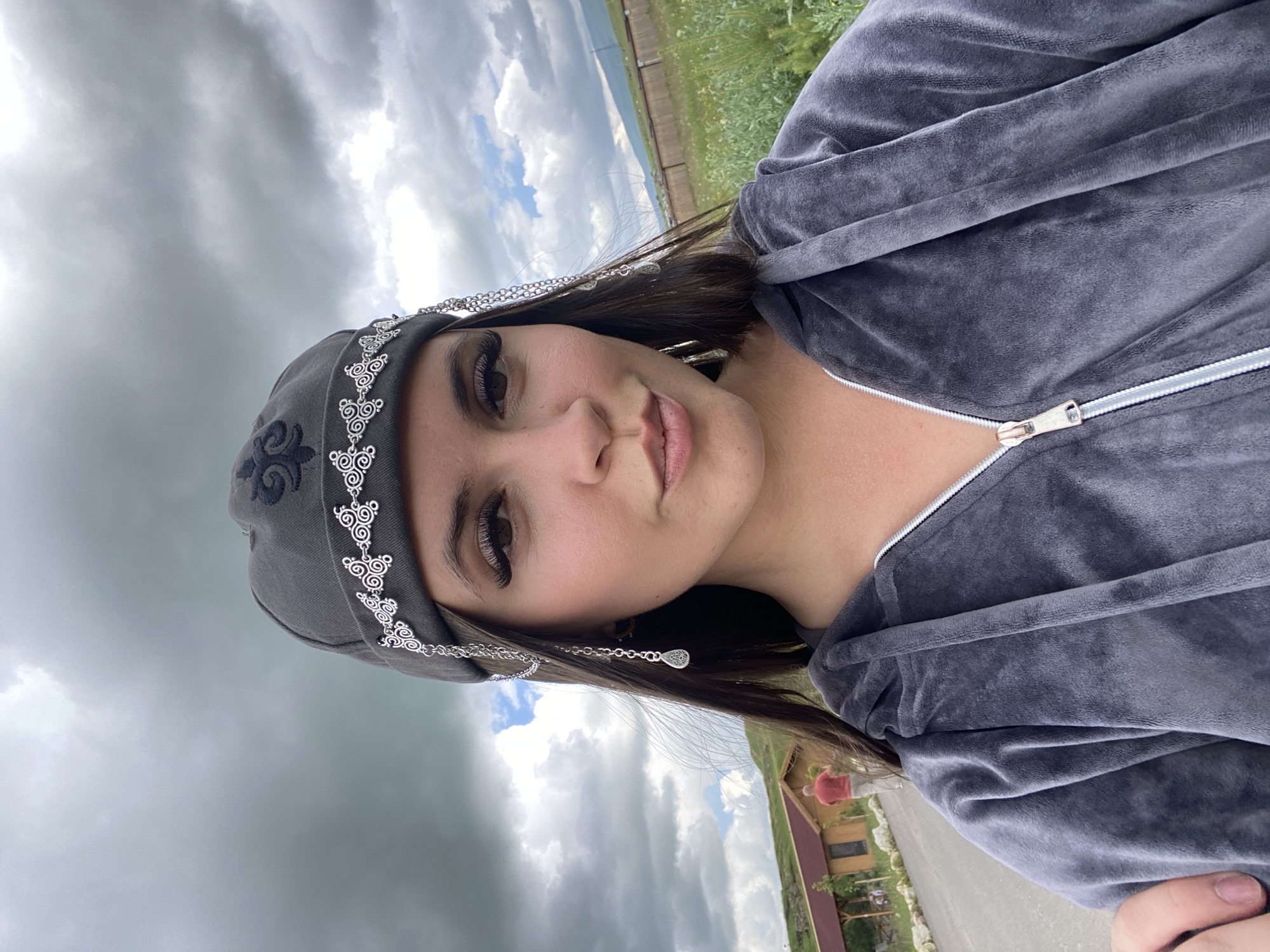 A selfie is taken outdoors in front of a cloudy sky. The subject is a young woman wearing a gray velour tracksuit and a gray beanie adorned with silver embellishments and dangling chains with tear-drop shaped charms. Her face is partially visible, and she has long dark hair. In the background, there is a road, a wooden building with a red roof, a person walking away from the camera, a fenced-off field with greenery, and a distant hillside. The location is identified as Tsalkita, Russia.
