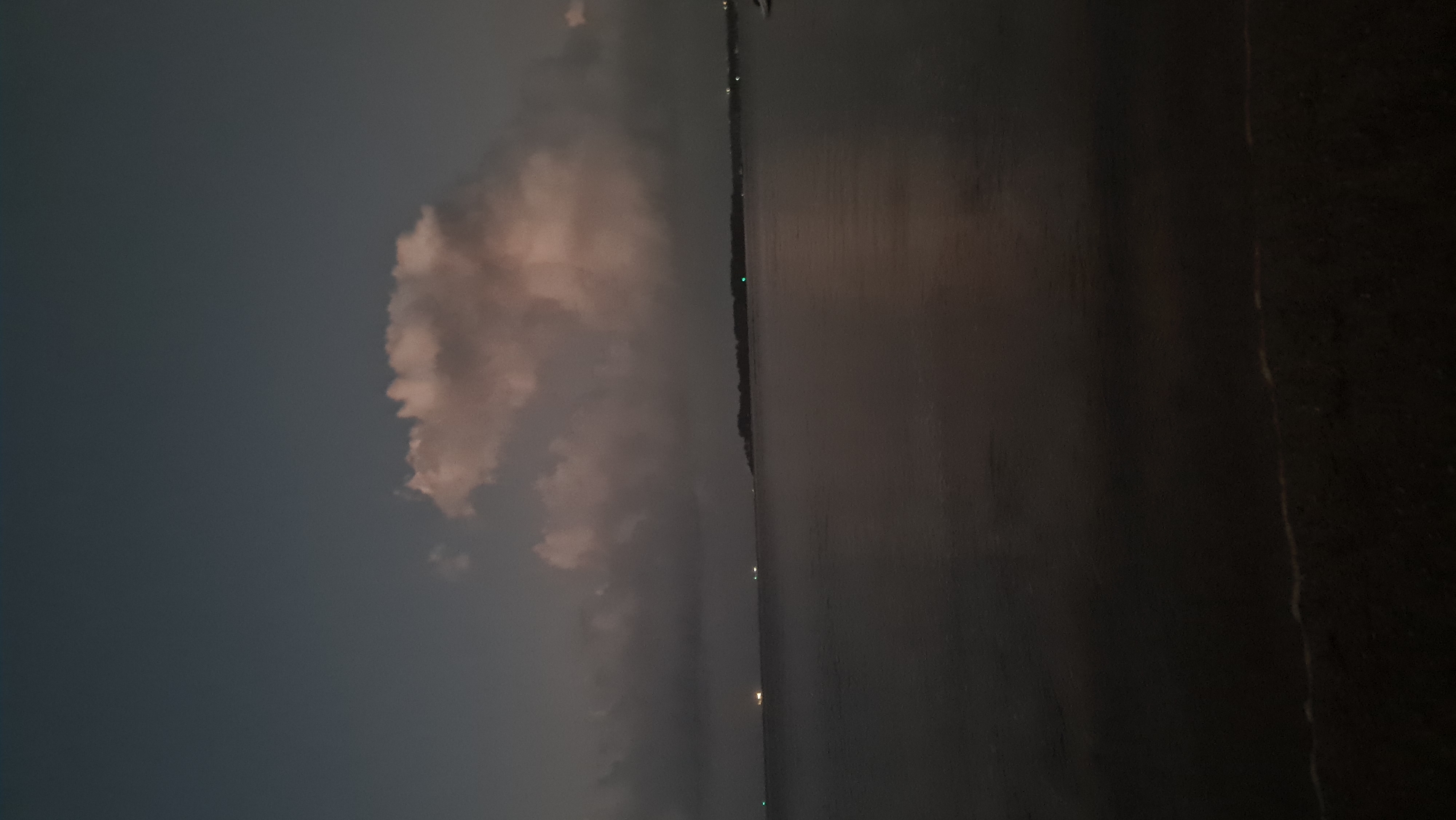 The image depicts a wide-angle view of a body of water, likely the ocean, at dusk or dawn. The sky is a muted blue-gray, with several large clouds that are tinged with pink and orange, suggesting the last or first light of the sun. There are distant lights visible on the horizon, which could indicate boats or a coastline with buildings. The water is dark and appears calm, reflecting the subtle colors of the sky. In the foreground, there is a dark strip of land, possibly a beach or shoreline, with very little detail visible due to the low light conditions. The overall impression is one of a tranquil and somewhat subdued natural scene. Without specific landmarks or identifying features, it is not possible to pinpoint the exact location within Denpasar, Indonesia.