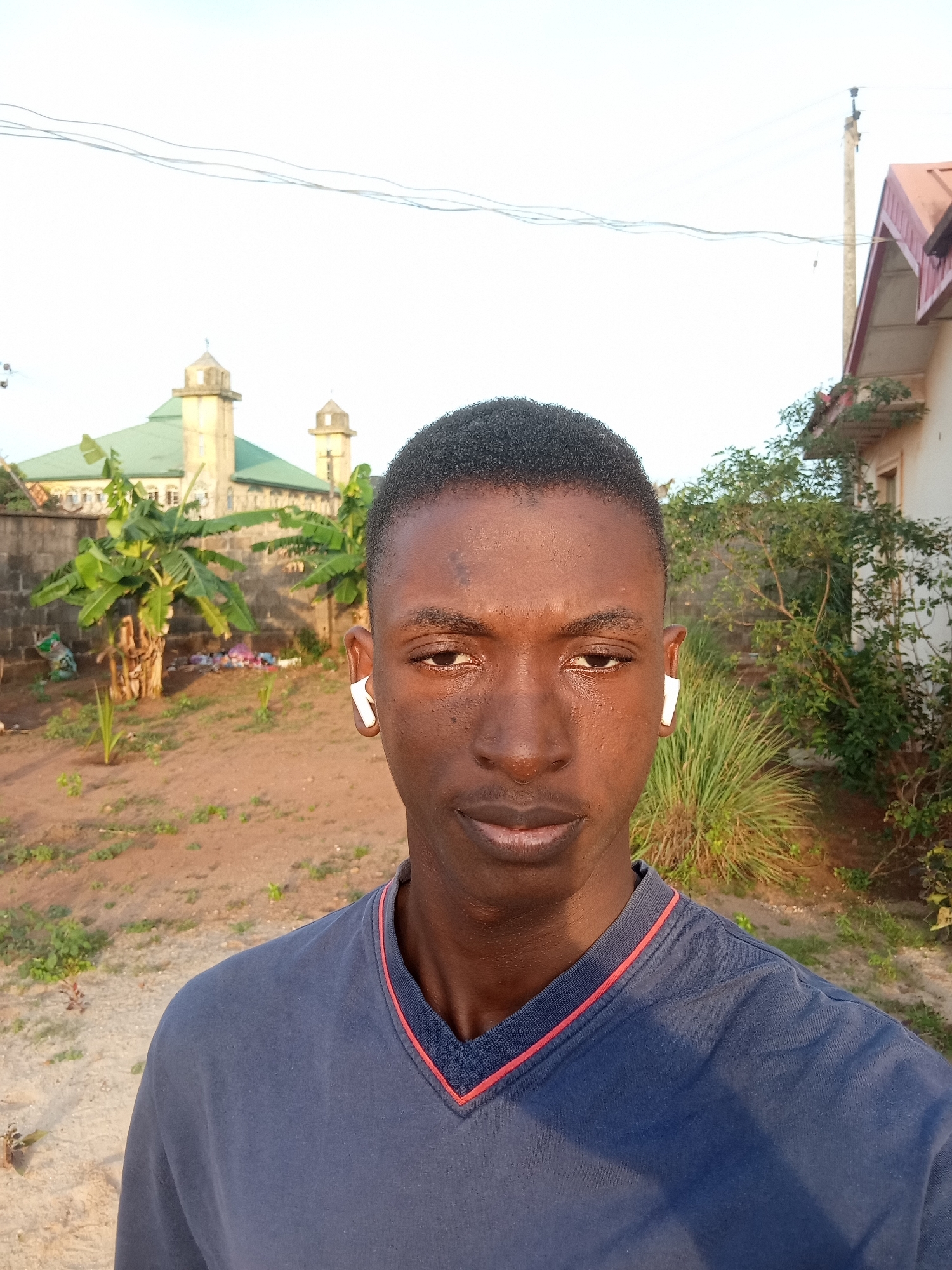 A medium shot shows a young Black man wearing a dark blue V-neck t-shirt and white wireless earbuds standing in a yard. Behind him is a building with a red roof on the right and a green-roofed building with twin towers on the left. Banana trees and various other plants are scattered in the yard. The location is Ikorodu, Nigeria.