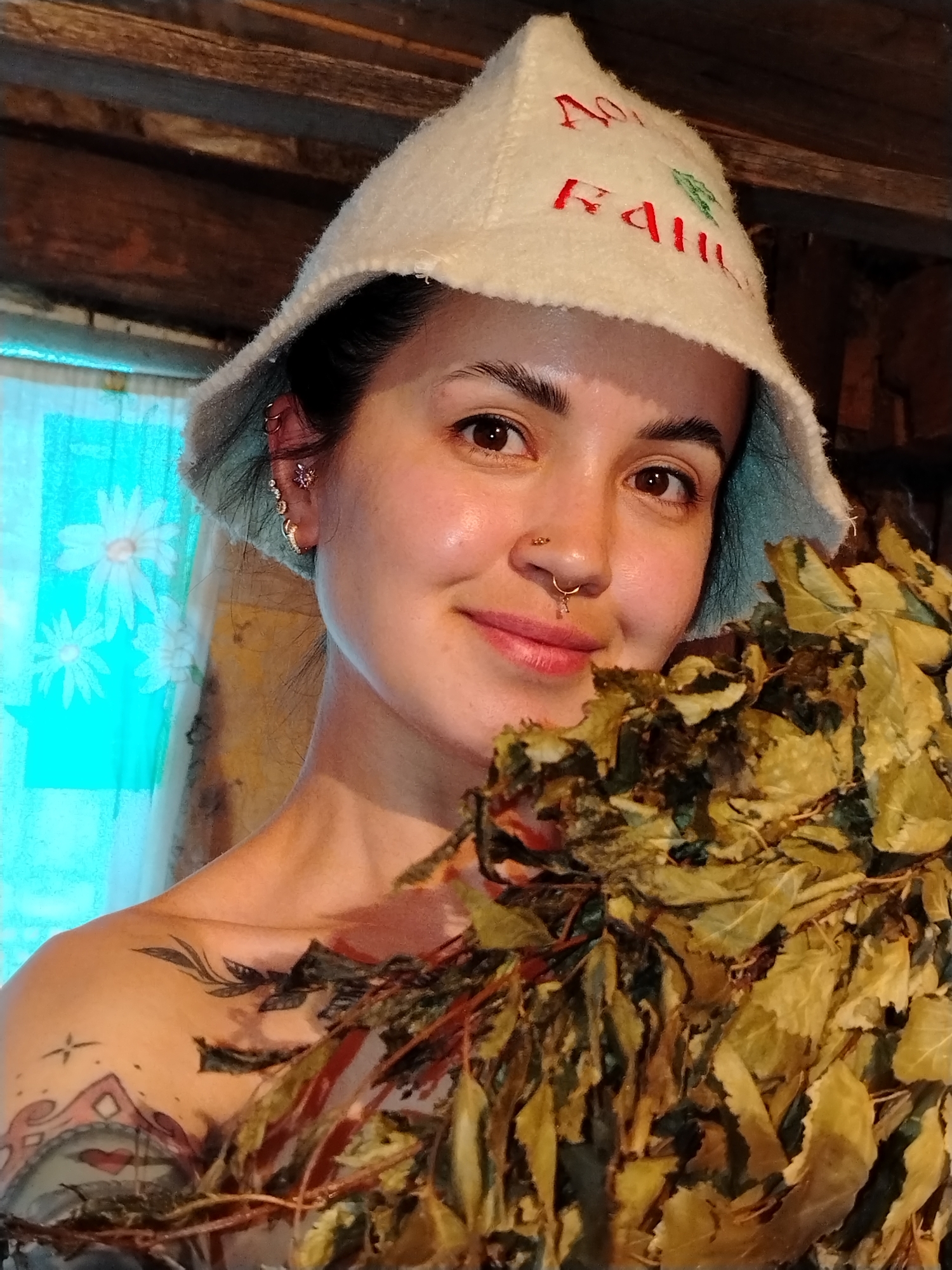 A woman is holding a bundle of dried leaves, possibly birch branches, in front of her. She is wearing a light-colored felt hat with lettering in red that might be Russian. The background appears to be a sauna or bathhouse interior with wooden walls and a floral patterned curtain. She has multiple piercings in her ear and nose and visible tattoos on her chest and shoulder. Given the hat, the location is likely Гожан, Russia.