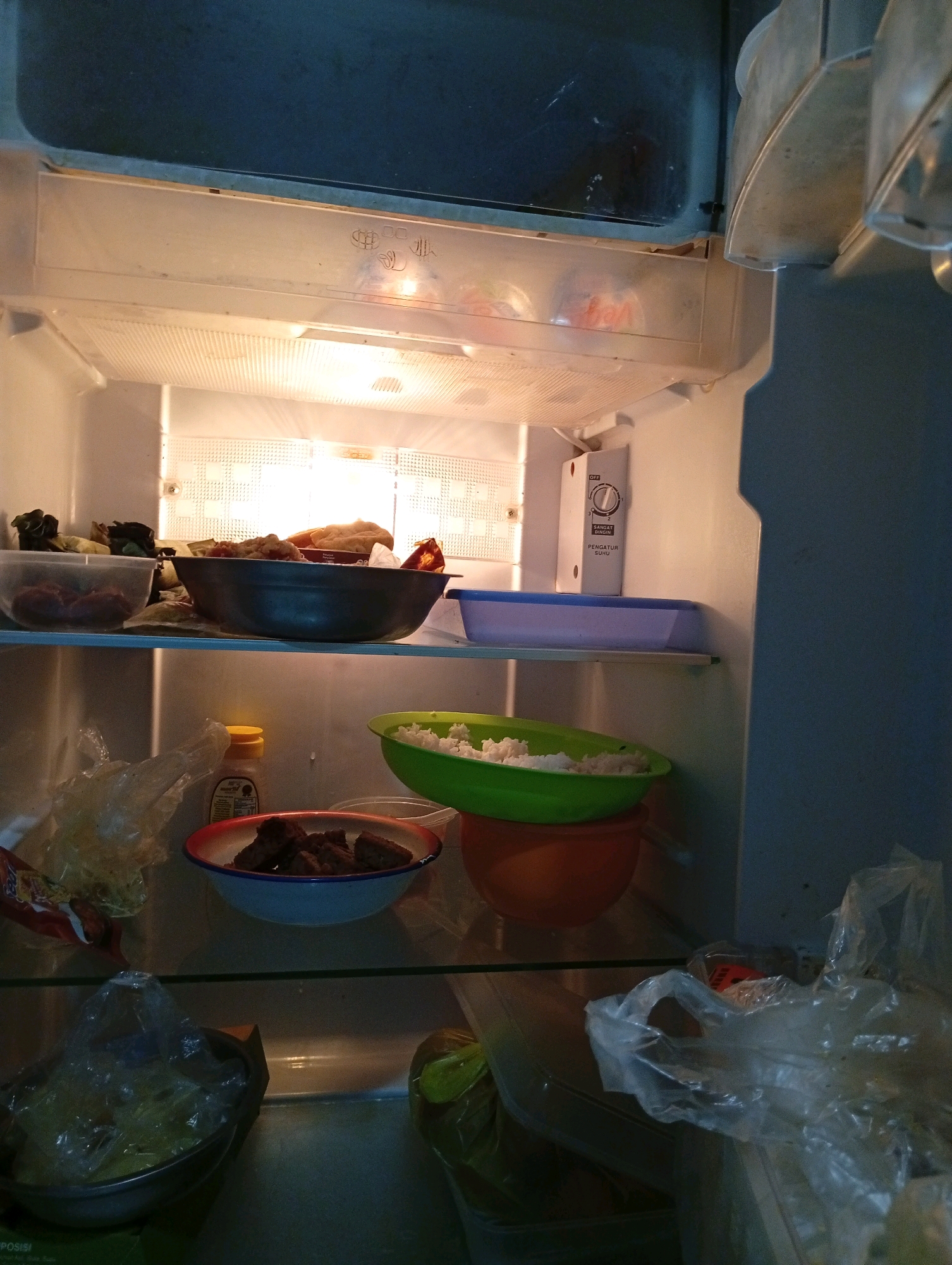 The image shows the inside of an open refrigerator, dimly lit by its internal light. On the top shelf, there are various food items, including what appears to be cooked meat and some leafy vegetables in plastic containers. A large dark bowl containing food is centrally placed on this shelf. Below this, on the next shelf, are two plastic containers: one is a light blue rectangular container, and to its right, a green bowl filled with white rice sits atop an orange bowl. Further down, on the lowest visible shelf, a red and white bowl holds several pieces of cooked meat. To the left, a partially obscured clear plastic bag contains more food items. In the foreground, another clear plastic bag with a handle is visible, and to its right, a large plastic bag also contains food. The overall environment is cluttered with food items and packaging, suggesting preparation or storage of meals. The background is the interior wall of the refrigerator, showing its construction and a temperature control dial with Indonesian text, indicating the location is likely within Indonesia, potentially in a place like Kebondalem.