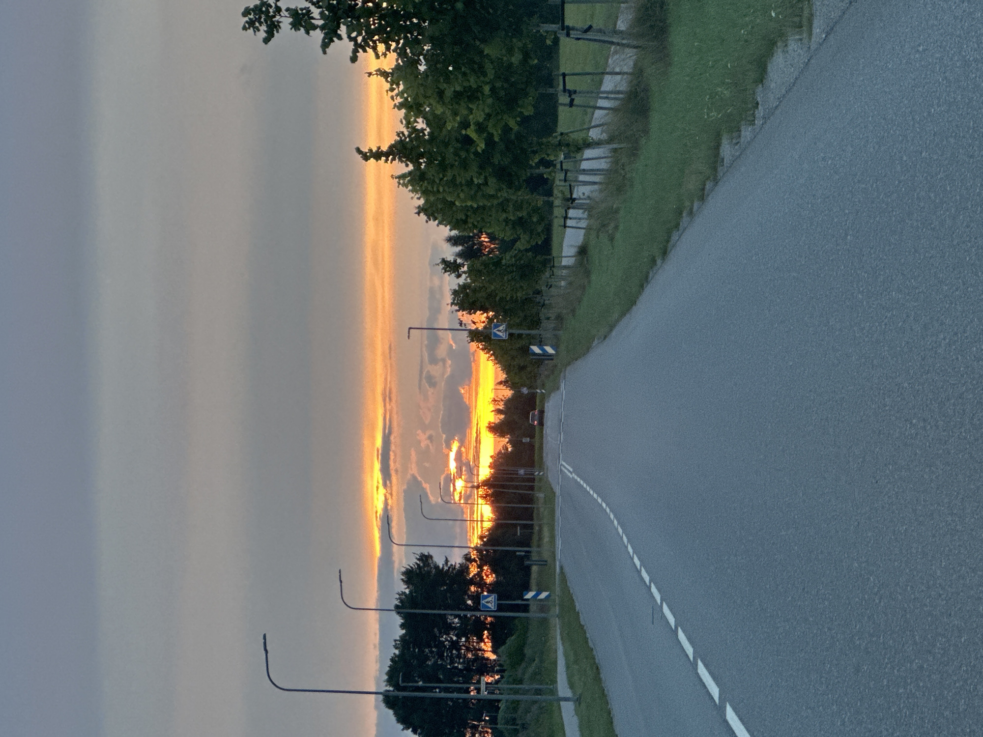The image depicts a rural road in Tiskre, Estonia, during sunset. The sky is filled with a dramatic display of orange and yellow hues, with scattered clouds casting shadows. The road, paved with asphalt, stretches towards the horizon, marked by dashed white lines on the left side. Several streetlights line the left side of the road, their modern designs silhouetted against the sky. On the right side, a grassy verge borders the road, with a row of young trees, each supported by stakes, planted along its edge. Beyond the trees, a fence is visible. A single car is seen in the distance on the road, its presence minimal against the vastness of the landscape and sky. The overall scene conveys a sense of tranquility and natural beauty at the end of the day.