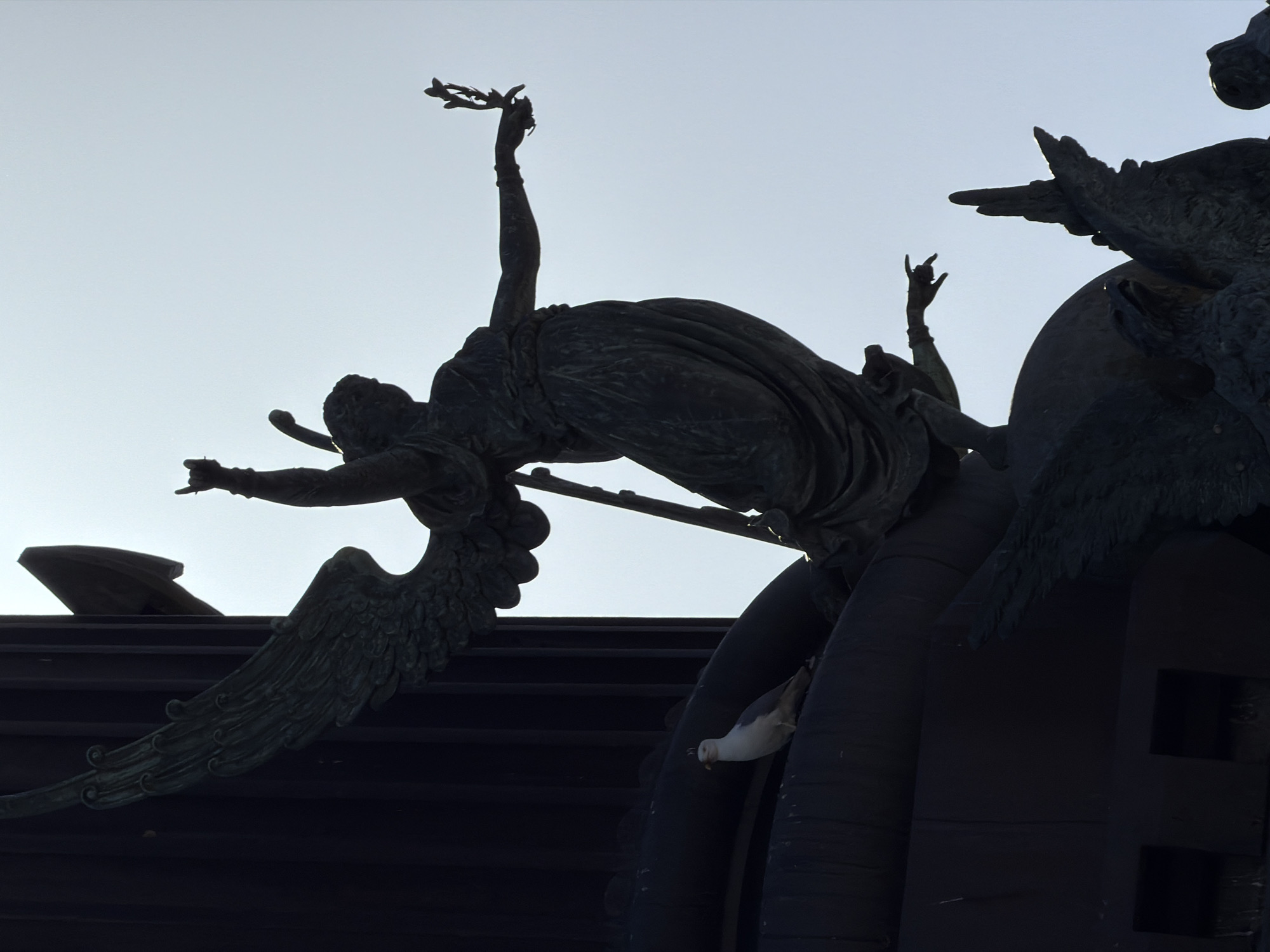 The image shows the Monument to Columbus in Barcelona, Spain, at the end of Las Ramblas. A bronze sculpture of an angel, part of the monument, is silhouetted against the sky. The angel has one arm raised, holding a laurel branch, and the other arm bent with the hand pointing. A seagull is perched on a dark, curved surface in the foreground. The monument is made of dark bronze, and the sky is a pale, hazy blue.