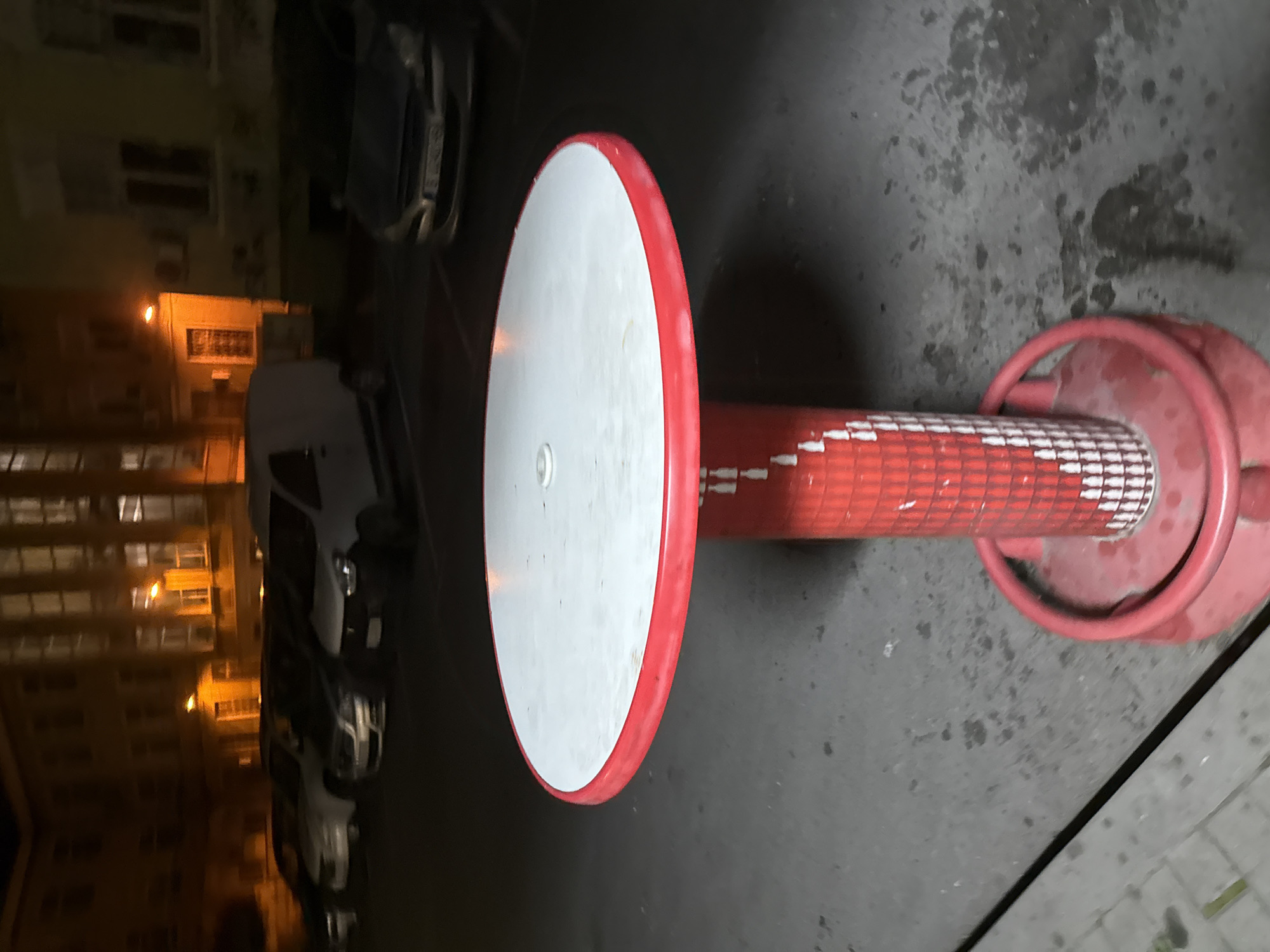 The image shows an outdoor scene at night in what appears to be Varna, Bulgaria, evidenced by the architecture in the background. The main subject is a red and white, round, fixed table-like structure on a gray paved surface. The table has a white top with a red rim. A red cylindrical post connects the table top to a round base on the ground. The post has a pattern of white triangles pointing downwards. The background features blurred buildings with windows lit up by yellow street lights. Several cars are parked along the street. The scene appears to be a public space, possibly a recreational area or park. No people are visible.