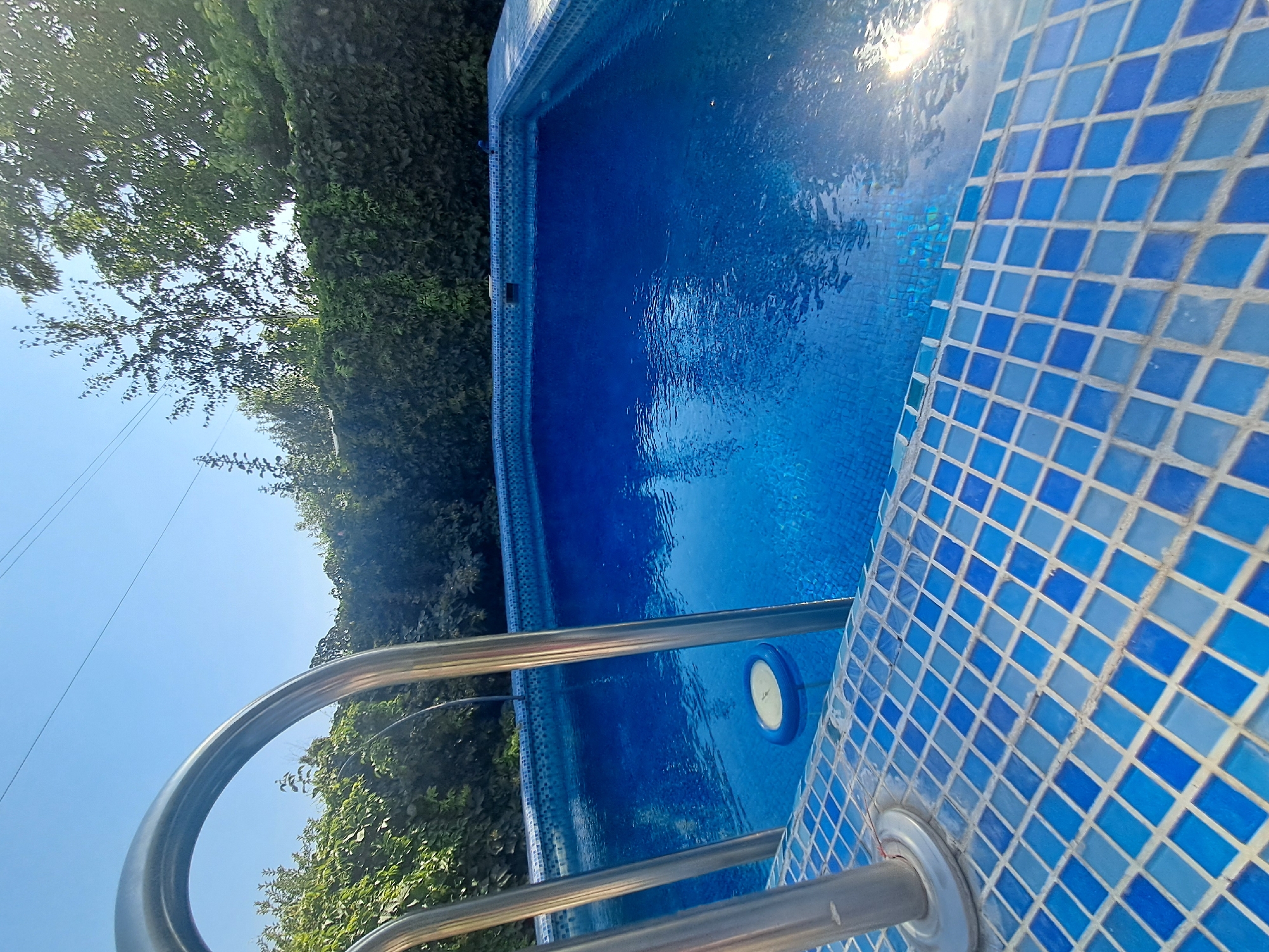 The image depicts a close-up view of a swimming pool, with a focus on the tiled edge and the clear blue water. A metallic handrail is visible in the foreground, curved and shiny, reflecting the sunlight. The pool is surrounded by lush green foliage and a clear blue sky. The sunlight glints off the water, creating a sparkling effect. The tiles lining the pool are a mosaic of different shades of blue, adding a decorative element to the scene. A circular pool light is submerged in the water. The overall impression is one of a tranquil and inviting outdoor setting, likely on a sunny day. The scene suggests a moment of relaxation and enjoyment, with the pool ready for use. No people are visible in the image. The location is identified as Shymkent, Kazakhstan, which implies a warm climate suitable for swimming. The time of day appears to be daytime, given the bright sunlight. There is no visible text in the image.