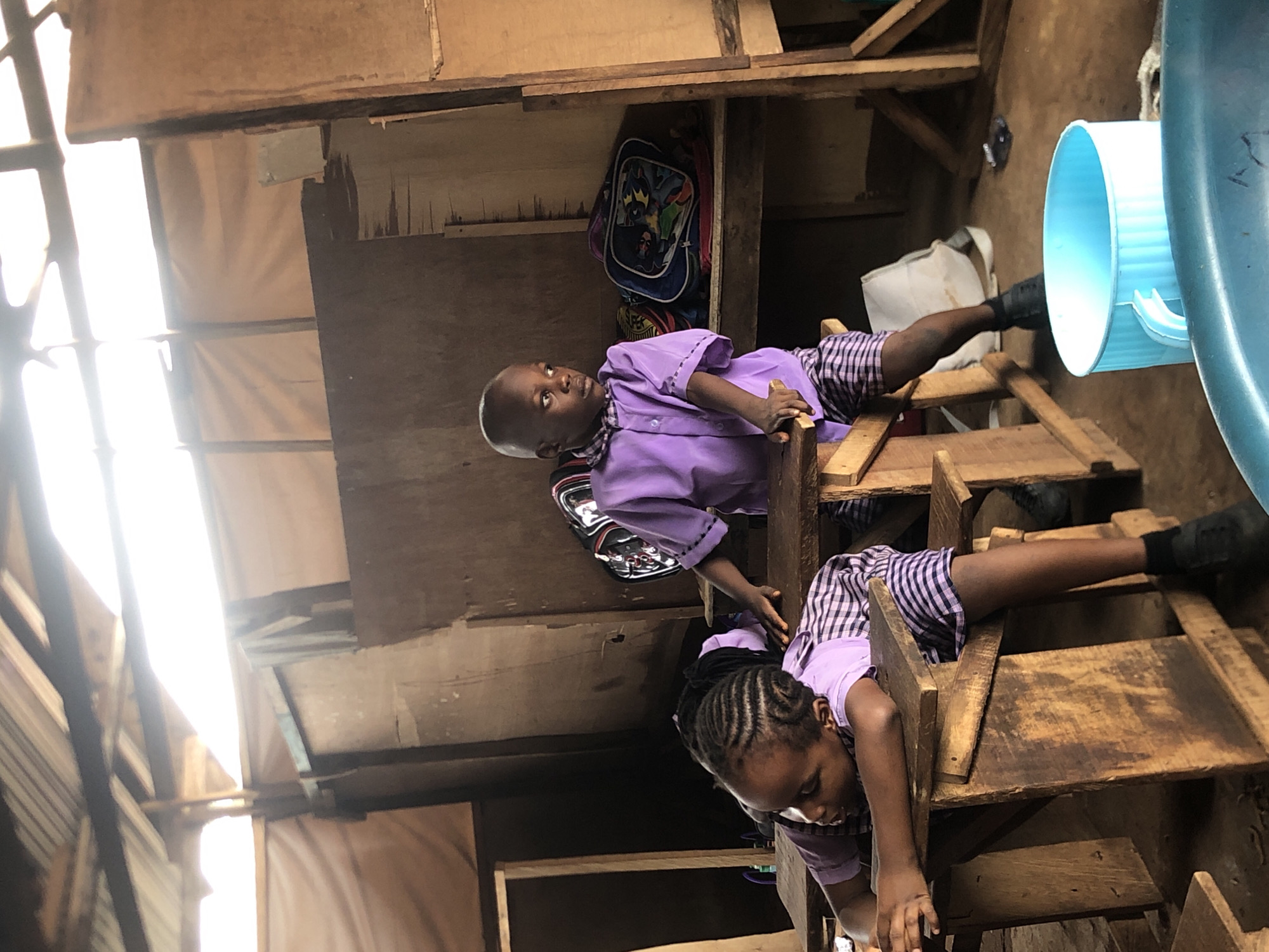 In a dimly lit, makeshift classroom structure in Ukwu-Abwa, Nigeria, two young children are observed. The structure appears to be constructed from wood and corrugated metal roofing, with brown fabric lining some of the walls. Sunlight streams in through gaps in the roofing, creating bright spots.
In the foreground, a child with intricately braided hair is slumped over a wooden desk, their head resting on their arms. They are wearing a purple collared shirt and a plaid skirt. To the right, a bald-headed child in a similar purple collared shirt and plaid shorts stands next to a wooden desk, looking to the right with a contemplative expression. Behind this child, a blue backpack with cartoon characters is visible on a shelf. A large, light blue plastic bucket is in the lower right corner of the frame. The overall atmosphere suggests a rural or impoverished educational setting.