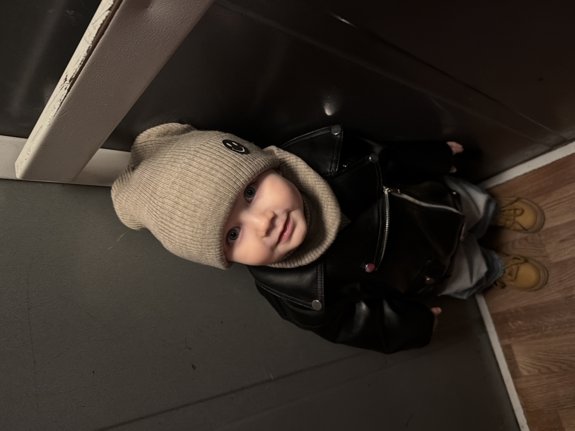 A baby wearing a tan knit hat with a smiley face, a black leather jacket, and yellow boots is positioned in the corner of a room, possibly in Chaikovsky, Russia. The walls are gray, and the floor is wooden. A white trim or molding is visible along the floor and on the left wall. The baby is looking up at the camera.