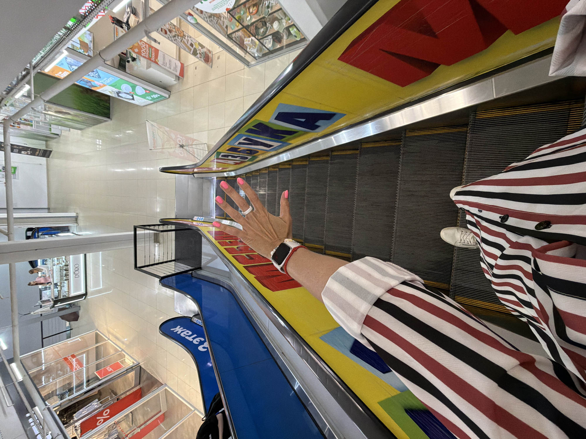 The image shows a first-person perspective of a person on an escalator in a shopping mall. The person is wearing a striped shirt and white sneakers. Their left hand, with bright pink nail polish and a ring, is visible near the top of the escalator. The escalator has yellow and red lettering on its side, which appears to spell out "АЗБУКА" (Azbuka, meaning "Alphabet" in Russian).
The shopping mall environment features other escalators, glass-fronted shops with red sale signs, and displays of merchandise. The lighting suggests an indoor setting during the day. The overall scene is typical of a retail complex. The specific location in Birobidzhan, Russia, cannot be confirmed from the visual information alone.