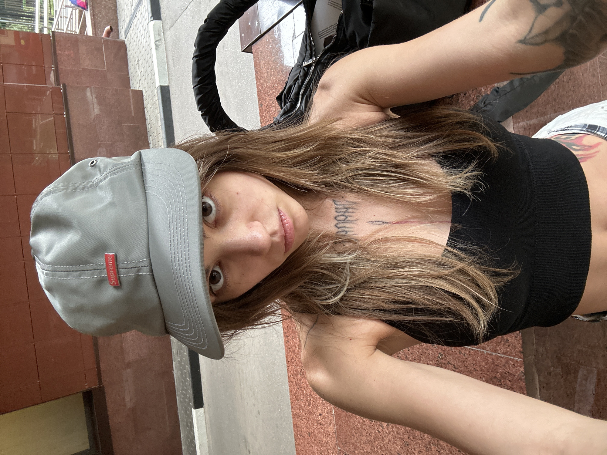 A young woman wearing a grey Supreme baseball cap and a black tank top, with visible tattoos on her chest and arm, is taking a selfie outdoors. She is seated on a tiered, reddish-brown stone surface, possibly a public bench or steps. Behind her, the facade of a building with reddish-brown stone tiles and a dark horizontal stripe is visible. To her right, part of a black bag with a prominent curved handle is in the frame. The ground in front of her is paved with reddish-brown and grey tiles. The context suggests a location in a city, potentially Moscow, Russia, given the architectural style and paved surroundings.