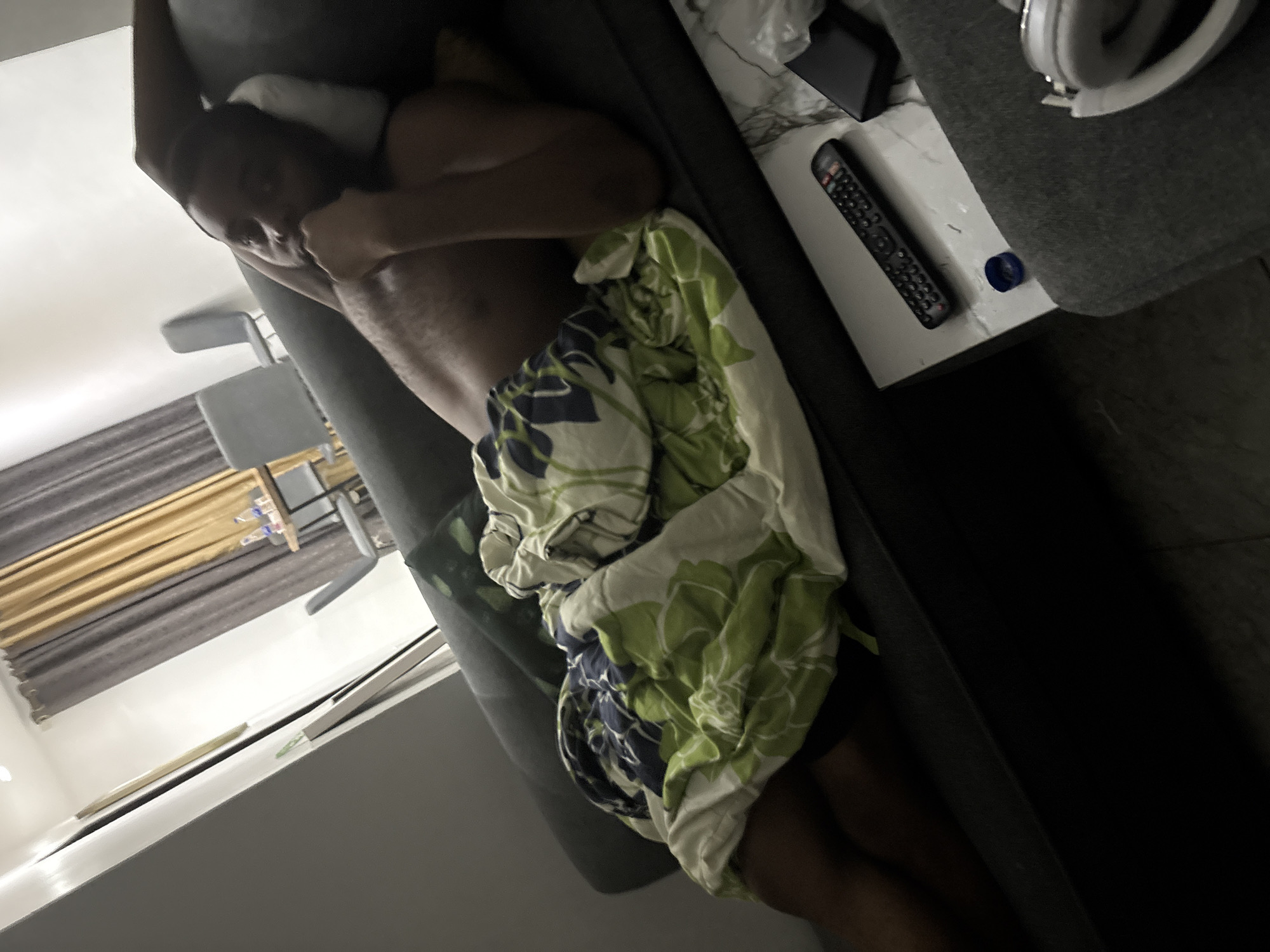 The image captures an indoor scene, likely a living room or bedroom, featuring a person relaxing on a dark gray couch. The person, appearing to be of African descent, is lying on their back with their head resting on the armrest or a pillow, looking towards the viewer with a relaxed expression. They are shirtless, and their lower body is partially covered by a colorful blanket adorned with green, white, and navy blue floral or leaf patterns.
Adjacent to the couch, a light-colored side table with a subtle marbled pattern holds several items. A black remote control, white over-ear headphones, and a small blue plastic cap are visible on this surface. The room's lighting is dim, suggesting it is either evening, night, or the room is intentionally dimly lit.
In the background, light brown or beige vertical blinds or curtains are visible, partially drawn, with darker gray curtains behind them. Further in the distance, two light-colored chairs with dark frames and a table are discernible. The overall activity depicted is one of comfort and rest. No visible text can be clearly read on any objects in the image. This scene provides a glimpse into a domestic setting, possibly in Iddo Tudunwada, Nigeria, given the provided location context.