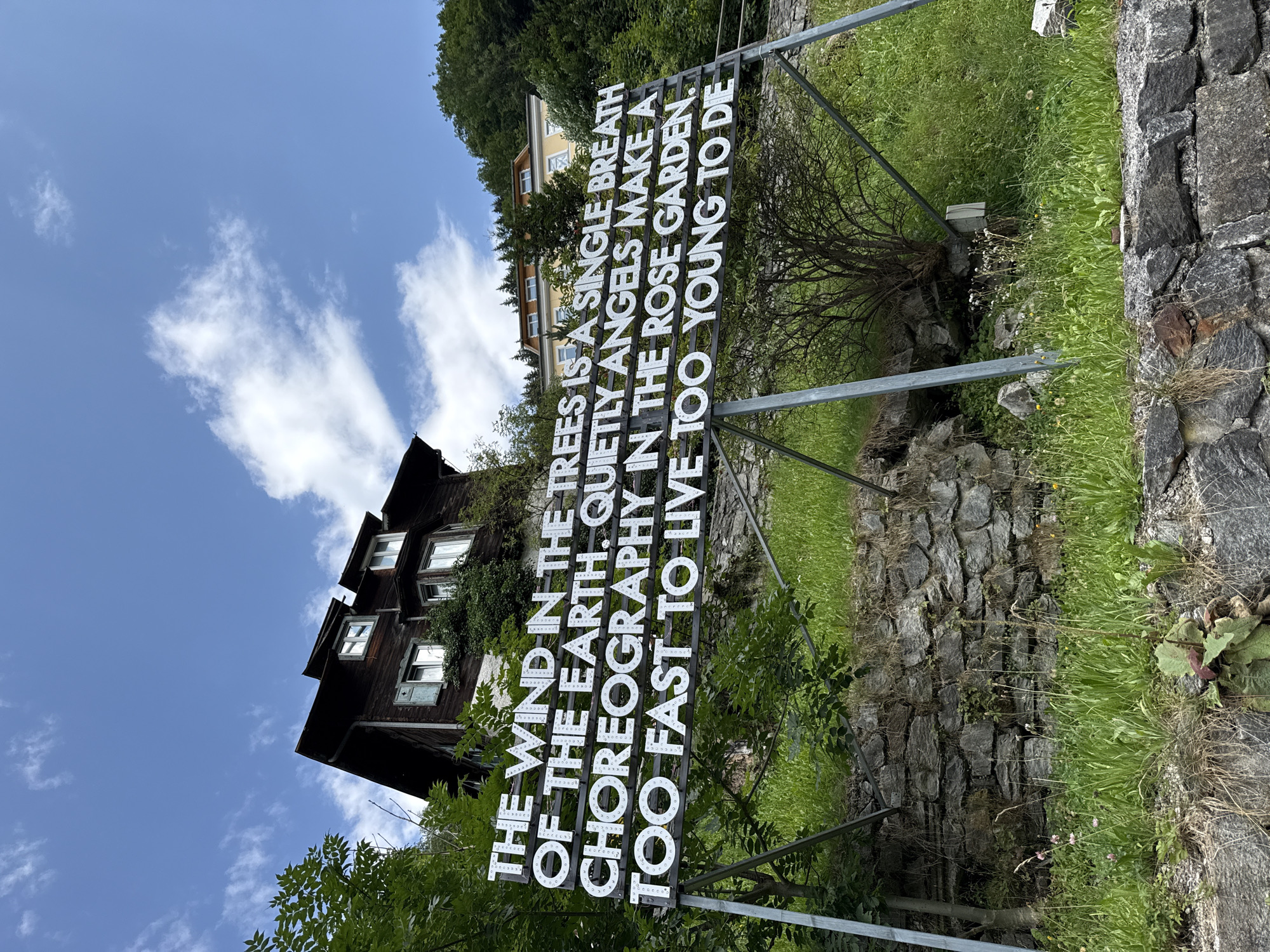 An outdoor, ground-level, wide-angle shot captures a large, white, illuminated sign with black text in the foreground. The sign reads: "THE WIND IN THE TREES IS A SINGLE BREATH OF THE EARTH. QUIETLY ANGELS MAKE A CHOREOGRAPHY IN THE ROSE GARDEN. TOO FAST TO LIVE TOO YOUNG TO DIE." The sign is supported by a metal frame structure. Behind the sign, a steep, grassy slope is visible, punctuated by a stone retaining wall. Further up the slope, a dark, wooden, multi-story house with dormer windows sits nestled amongst lush green trees. A bright blue sky with scattered white clouds forms the backdrop. The location is identified as the art installation "Die Zeit wird nicht weniger" (Time does not get less) by Franz West, situated near the Steinerner Steg in Bad Gastein, Austria.