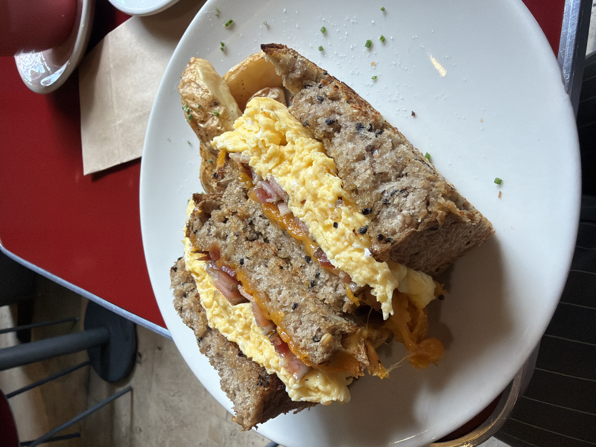A high-angle shot depicts a breakfast sandwich on a white plate, presumably at a dining establishment in Mexico City, Mexico. The sandwich, composed of two slices of seeded, whole-grain bread, contains scrambled eggs, cheese, and slices of ham. Roasted potato wedges are situated on the side. The sandwich is sprinkled with cut chives and salt. The plate rests on a table with a red surface. A partial view of a cup and saucer can be seen, along with a dark table leg in the background. The table is possibly located in an outdoor seating area given the hint of sunlight and view of the building.