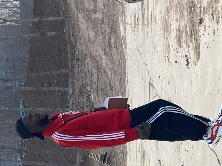 The image displays an adult male standing in profile, facing right, in an outdoor area of Zhongtun, China. The individual, who appears to be of African descent, wears a black baseball cap reversed, a red full-zip track jacket with three white stripes on each sleeve, and black track pants also featuring three white stripes down the sides. His footwear consists of multi-colored athletic shoes (white, orange, blue, black) with visible white socks. Sunglasses are suspended from the collar of his jacket, and a small black shoulder bag is worn across his torso. He holds a dark-colored smartphone with a yellow/green case in his left hand and a light brown folder containing white papers in his right hand.
The environment is characterized by dry, light brown, uneven ground with visible cracks and sparse debris. To the left of the individual, a yellow and black striped traffic cone is partially visible and tilted. The background features a large mound of dark brown soil, suggesting earthworks or construction activity. Several slender, bare-branched deciduous trees are scattered across the mid-ground and background, with some grey concrete blocks or pavers visible at the base of the soil mound. Behind the trees and soil mound, a multi-story building with a light grey or dull brown facade is visible.