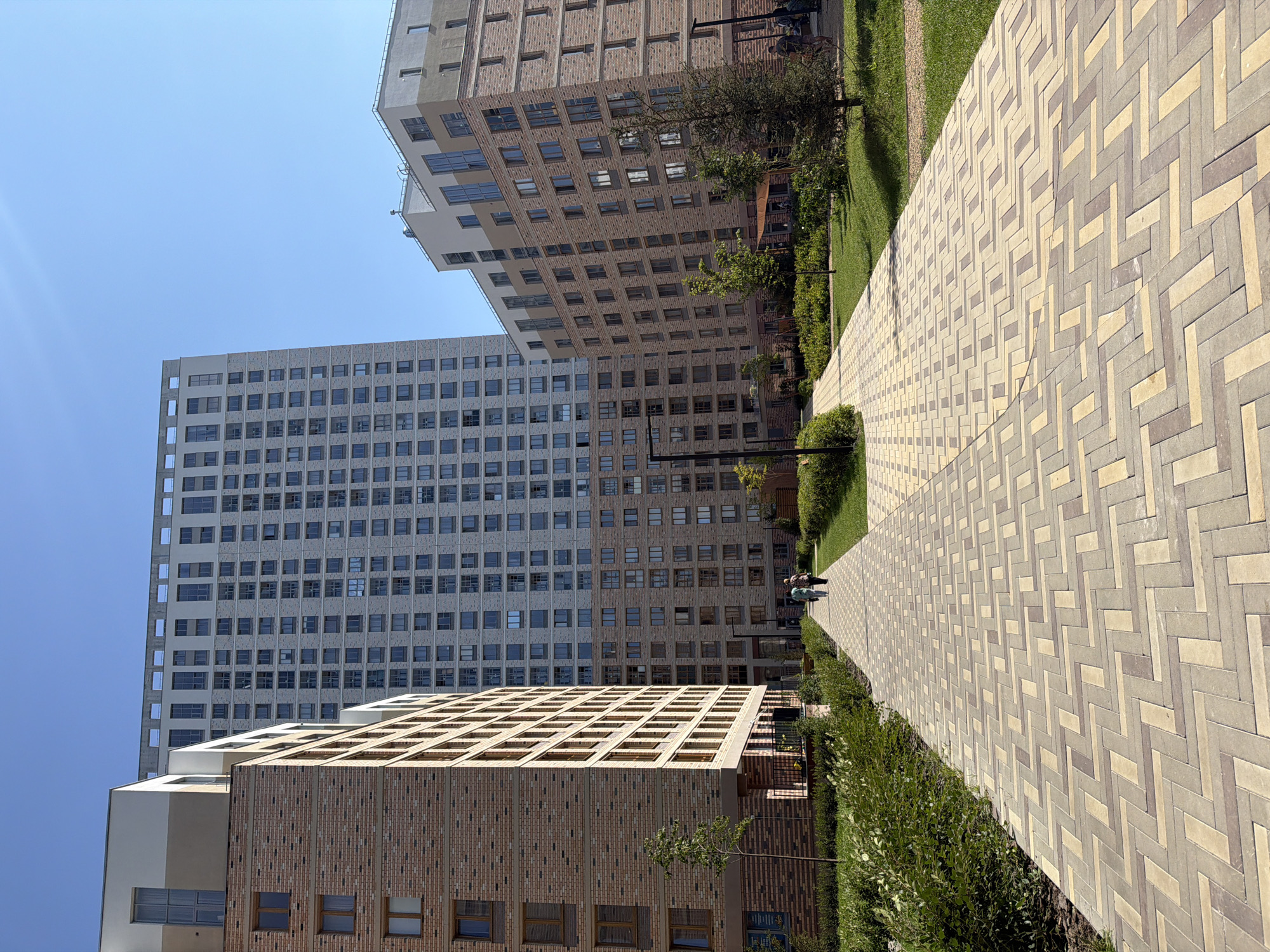 The image depicts a contemporary residential complex on a clear, sunny day. In the foreground, a paved walkway with a herringbone pattern made of tan, beige, and grey bricks leads towards a cluster of modern buildings. To the left, a brick building with multiple floors is visible, its facade composed of light brown bricks and rectangular windows. To the right, a section of a park area with manicured green grass and small trees is present.
Dominating the background is a very tall, slender building with a light gray facade and numerous square windows arranged in a grid. Adjacent to this tall building and slightly to the right, another multi-story building is visible, featuring a facade of red-brown bricks and a lighter colored top section with larger windows. Several people are walking away from the viewer on the paved pathway, dressed in casual clothing. The overall environment suggests a well-maintained urban residential area with a focus on modern architecture and green spaces. The architectural style and the presence of modern apartment buildings are consistent with developments in Yekaterinburg, Russia, a city known for its ongoing urban renewal and construction of new residential complexes.
