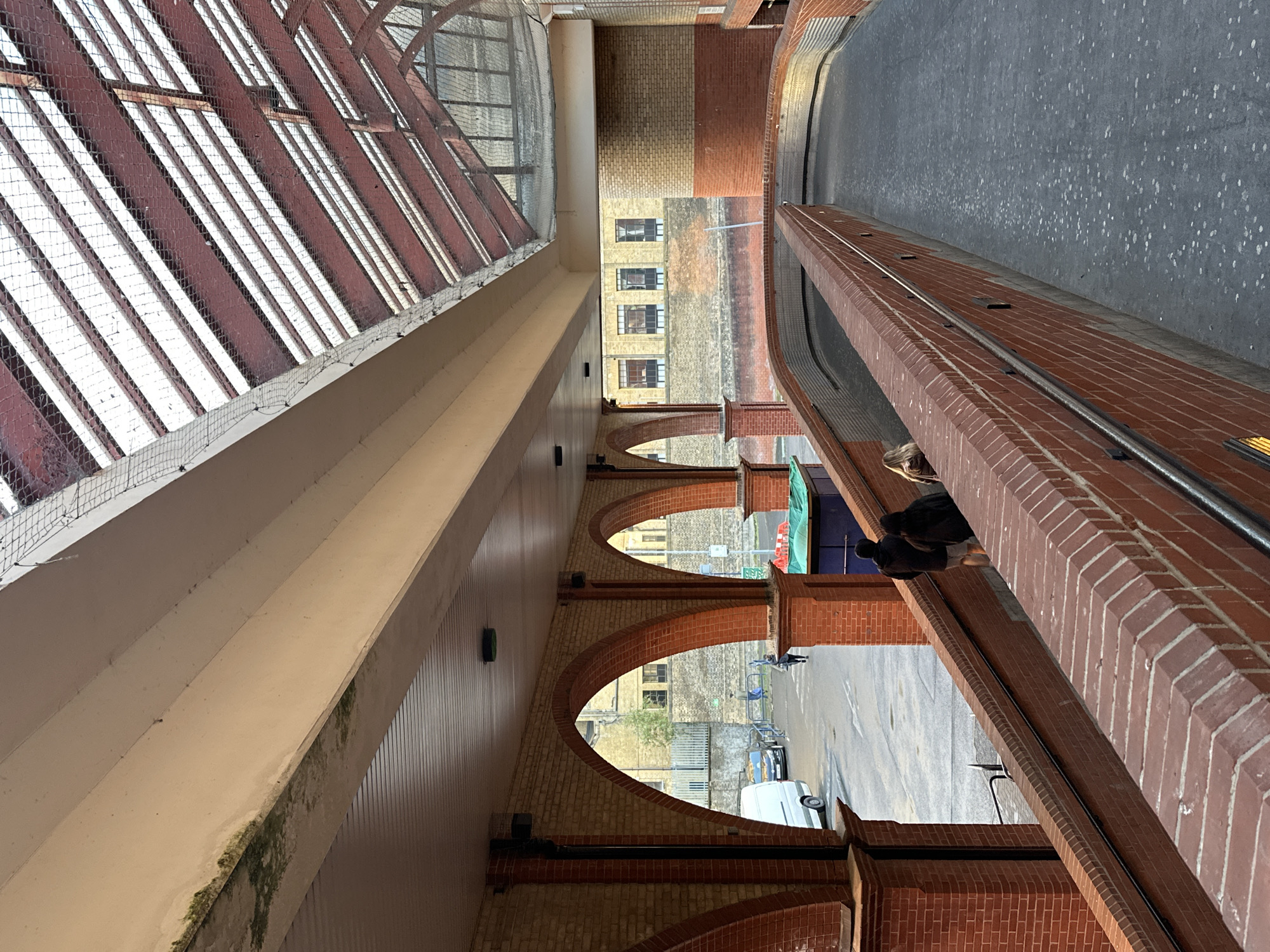 The image shows an elevated view of a walkway or bridge in what appears to be an urban setting. A large glass roof structure is visible on the left side of the frame, covered with a net, suggesting a possible architectural feature or public space. A series of brick arches line the path, offering glimpses into the streets below. In the distance, buildings with windows are visible. On the right side, a brick wall runs alongside the walkway, and people are seen standing beside it, their bodies partially obscured. A gray asphalt surface stretches out, and beyond it, more buildings can be seen. A white van is visible. The scene seems to be in daylight, but the time is not indicated.
Based on the architectural style and the overall urban landscape, it is highly probable that the location is in the city of London, United Kingdom, though exact confirmation of the specific location would require more context.