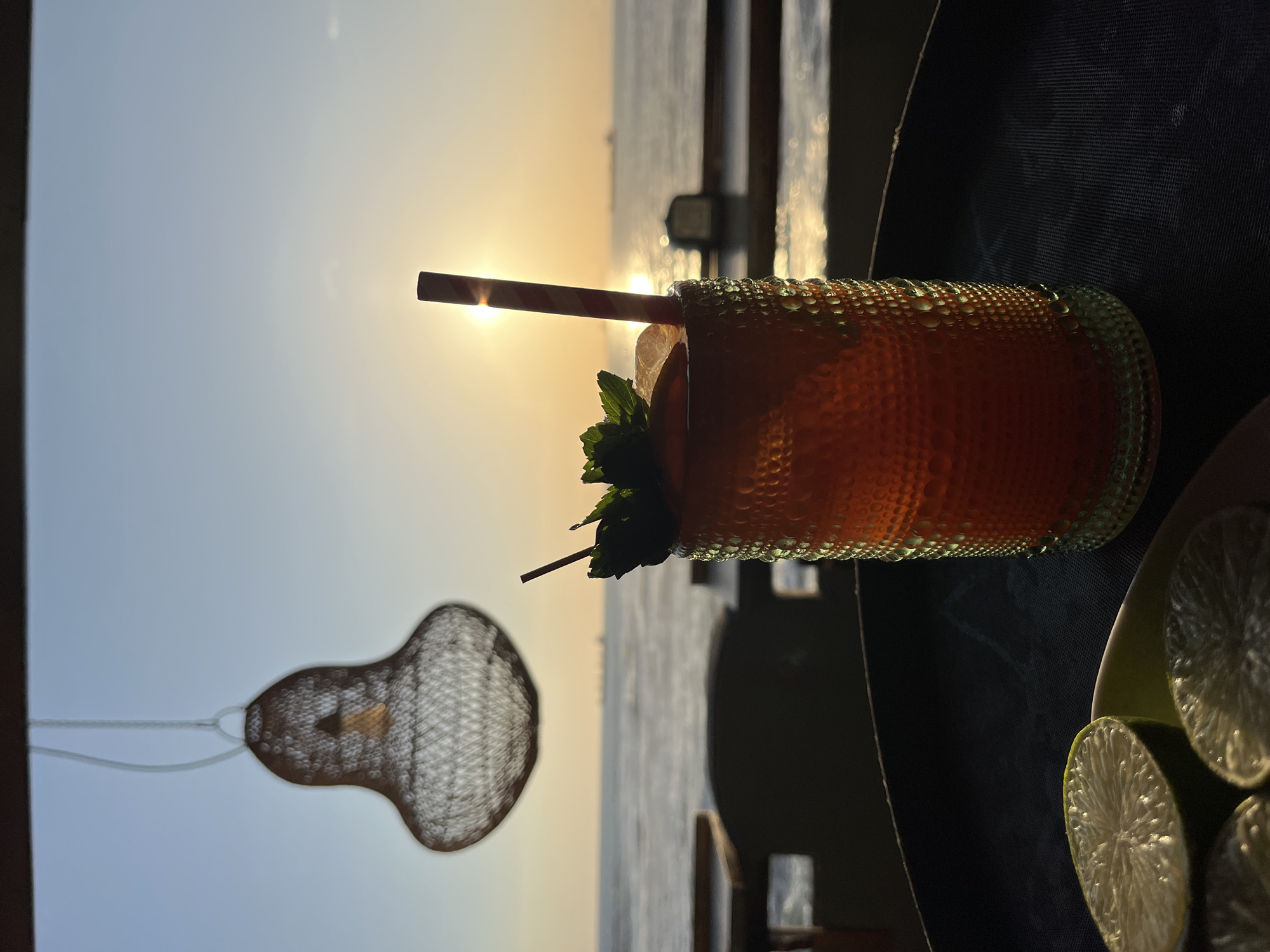 The image depicts a drink in a textured glass with a red and white striped straw and a sprig of mint, placed on a table alongside sliced limes. The background shows a body of water and the setting sun, suggesting an outdoor setting. A woven, pear-shaped light fixture hangs in the foreground. Given the description and clues in the image, it's likely this scene is in Arica, Chile, a coastal city known for its beaches and sunsets.