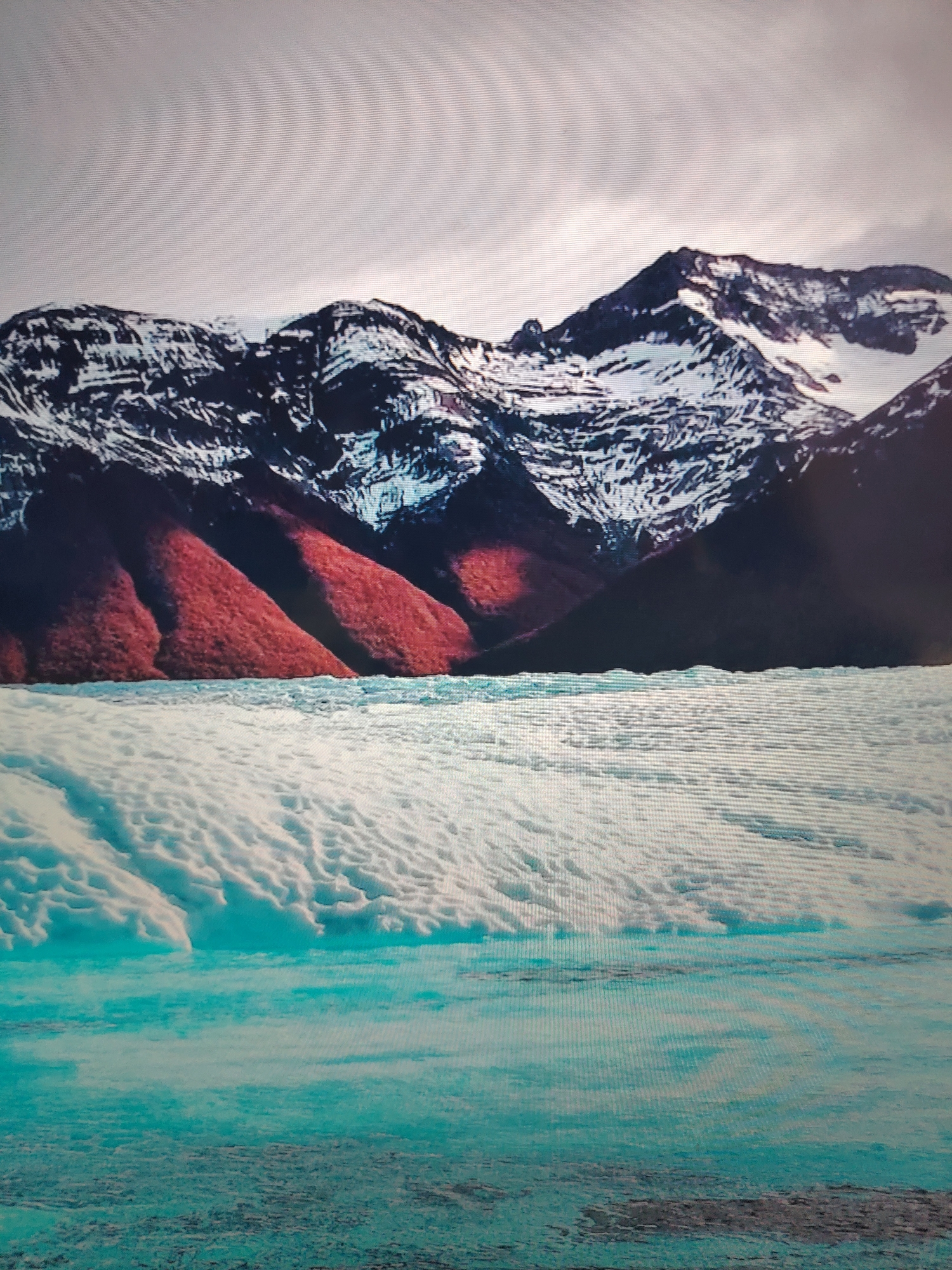 The image depicts a landscape featuring snow-capped mountains in the background, with lower slopes covered in dark, reddish-brown vegetation. In the foreground, there is a body of water, partially covered with ice. The water has a distinct turquoise hue. The sky is overcast with gray clouds. No people are visible. Location information is unknown.