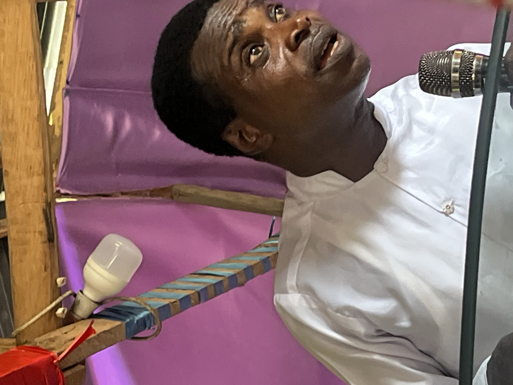 A man of dark complexion is prominently featured, seen from the chest up, actively engaged in speaking or singing into a microphone. He is dressed in a plain white collared shirt. His head is tilted slightly upwards and to the right, with his eyes wide open, gazing intently towards the top right of the frame, and his mouth slightly open, suggesting a moment of intense expression, perhaps passion, prayer, or vocal delivery. His brow is furrowed, indicating concentration or emotion.
In front of him, to his right, is a metallic microphone with a dark mesh head, positioned on a stand that is partially visible. The background is dominated by a vibrant purple fabric, likely serving as a curtain or backdrop, showing some creases. To the left of the man, a makeshift structure made of wooden planks and beams is visible. Attached to this structure is a white LED light bulb, illuminating the area. One of the wooden planks below the light bulb is painted with alternating blue and beige stripes. A small red piece of fabric or ribbon is also visible at the bottom left, attached to one of the wooden elements.
The setting appears to be an informal indoor or covered outdoor space, characteristic of a community gathering, possibly a religious service, public address, or performance in Iranla, Nigeria. The construction elements suggest a functional, handcrafted environment. No specific time of day can be determined, though the artificial light source suggests either evening or an interior space requiring illumination. No text is visible in the image.