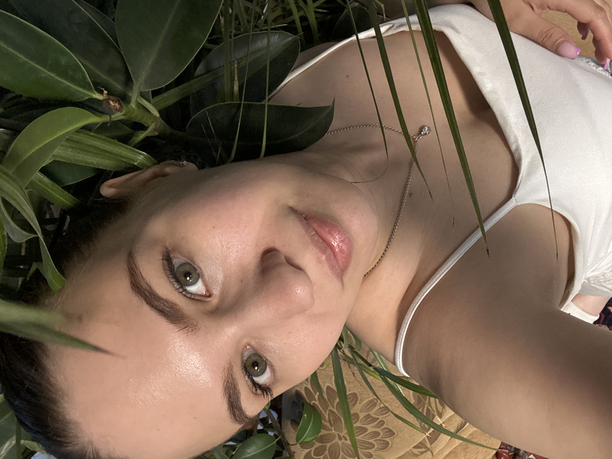A woman lies among green foliage. She wears a white tank top and a silver necklace with a small pendant. Her nails are painted a pale pink color. The background appears to be a patterned surface.
I am unable to provide the location in the city of Nizhnyaya Gar', Russia because there is nothing in the image to suggest it was taken there.