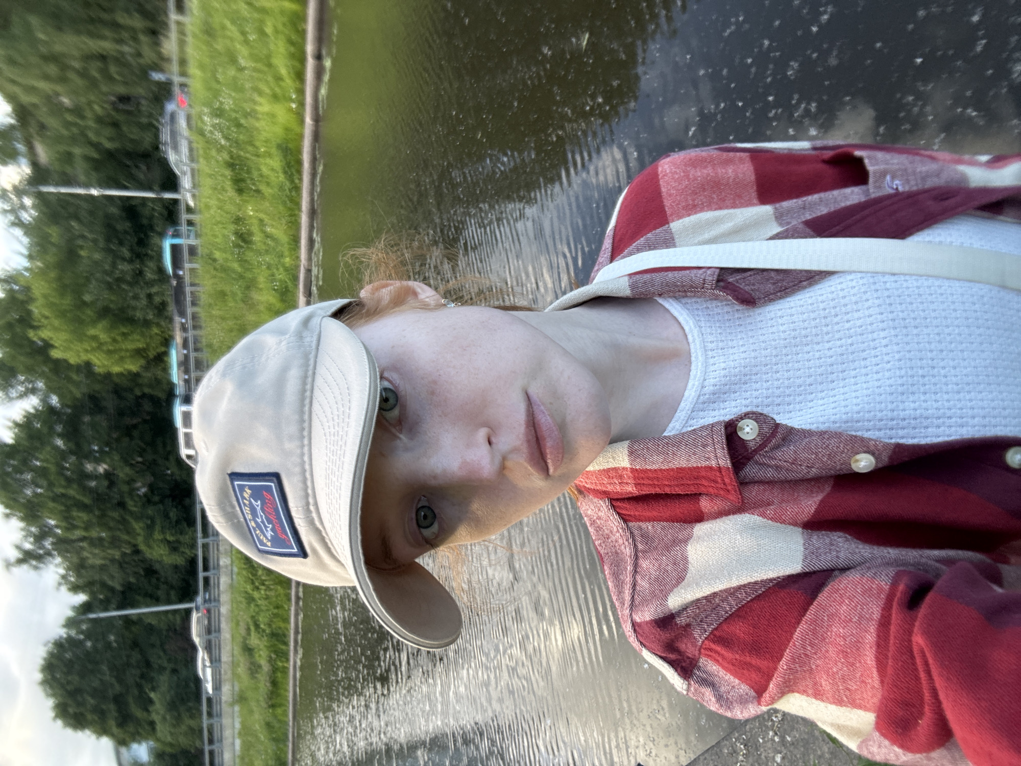 A young woman with red hair, fair skin, and green eyes is taking a selfie outdoors. She is wearing a light-colored baseball cap with a "Paul & Shark Yachting" logo, a red and cream plaid shirt, and a white waffle-knit t-shirt. A white strap from a bag is visible across her chest. She is standing on what appears to be a concrete embankment next to a body of water. In the background, there is a grassy slope with trees and a road with traffic, suggesting an urban or suburban environment. The image shows a cloudy sky. Given the presence of a canal and the architecture of the vehicles and road visible in the background, the location is likely within Saint Petersburg, Russia, possibly along one of the city's many canals or embankments.