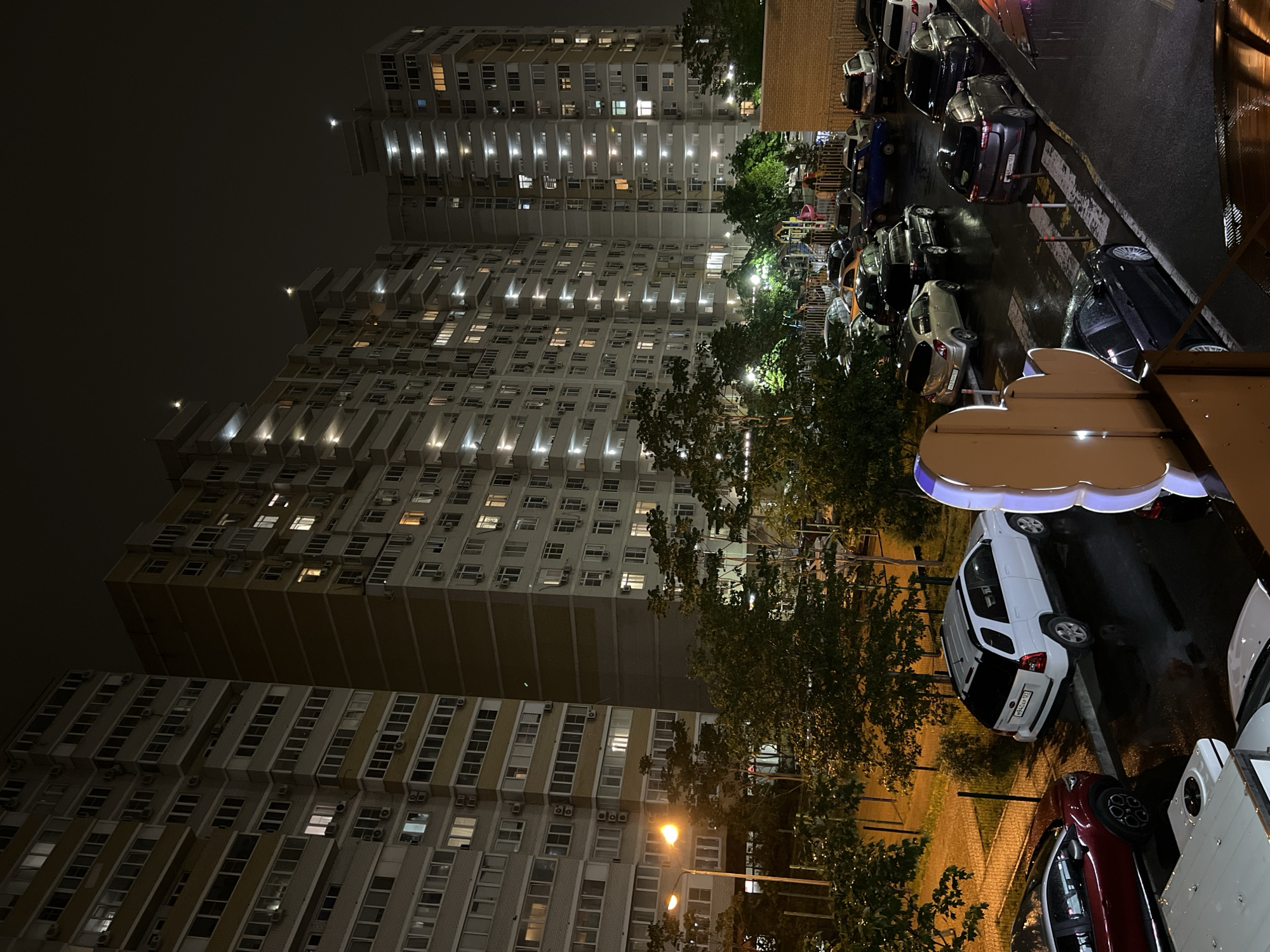 A low-angle shot captures a night scene in Krasnodar, Russia, emphasizing the architectural contrast and urban atmosphere. Several high-rise apartment buildings dominate the left side of the image, illuminated by numerous windows, indicating activity within. A street-level view on the right side reveals parked cars on a wet, reflective surface, suggesting recent rain. A leafy tree line separates the buildings from the street, and a street lamp provides additional illumination. The overall mood is that of an urban night scene, with the buildings casting long shadows. The exact location within Krasnodar is not discernible from the image alone.