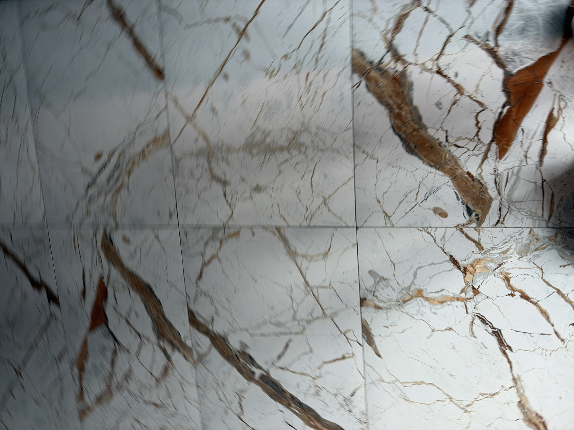 The image shows a close-up, blurry view of white marble tiles with brown and grey veining, likely a floor or wall. The lighting is dim and casts shadows, suggesting an indoor environment. There are no identifiable landmarks or clues to pinpoint a specific location within Moscow, Russia. The image is abstract and lacks clear subjects or actions.