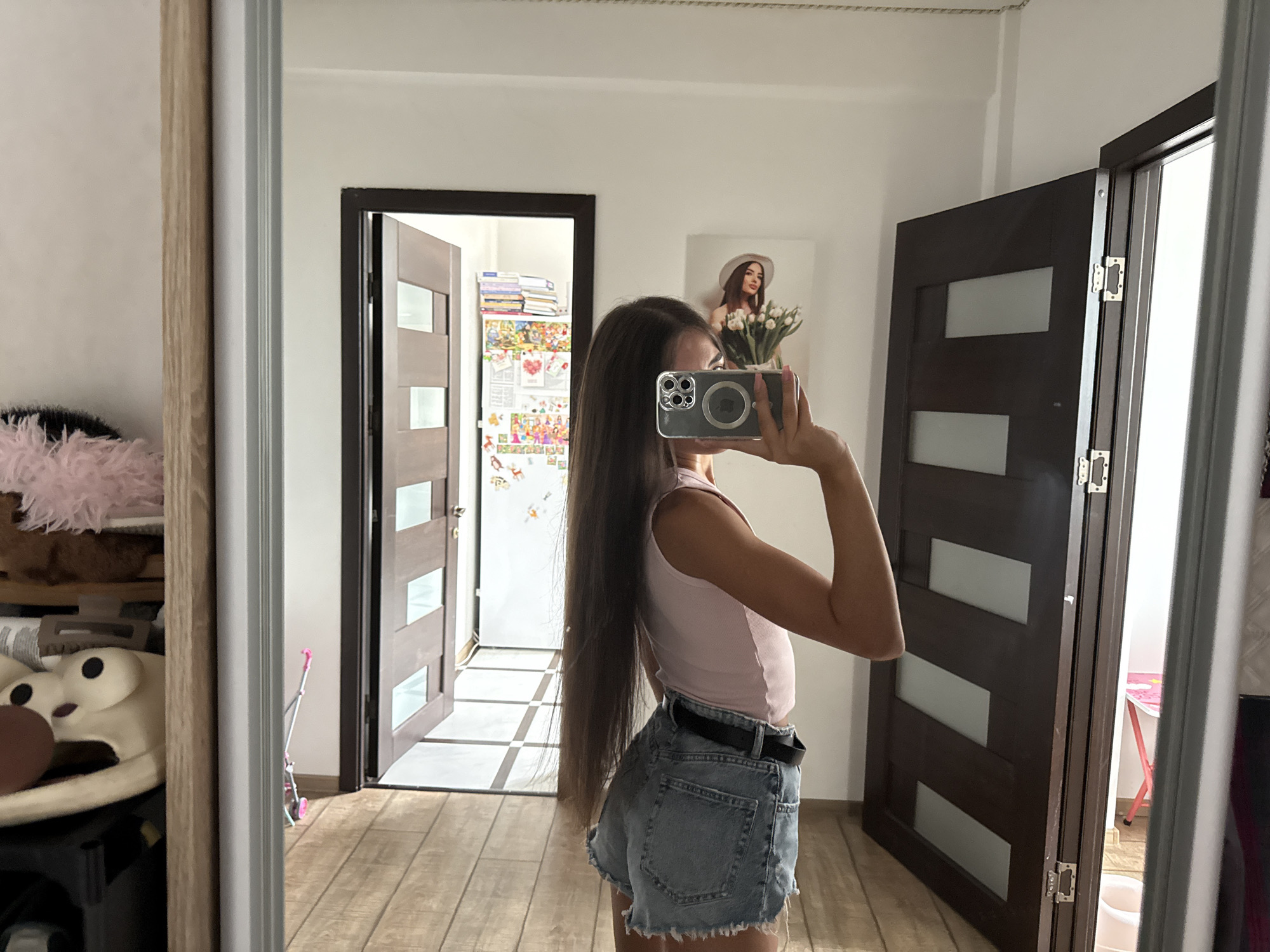 A young woman with long, dark hair is standing in an indoor space, taking a selfie in a mirror. She is wearing a pink sleeveless top and denim shorts with a black belt. Her right arm is raised, holding a smartphone to capture the reflection. In the background, two wooden doors are visible, one ajar revealing a hallway. To the left of the frame, a closet or shelving unit is partially visible with some stuffed toys. The room has wooden flooring and a light-colored ceiling. The overall impression is of a casual, personal moment captured within a residential setting. The location is identified as Chișinău, Moldova.