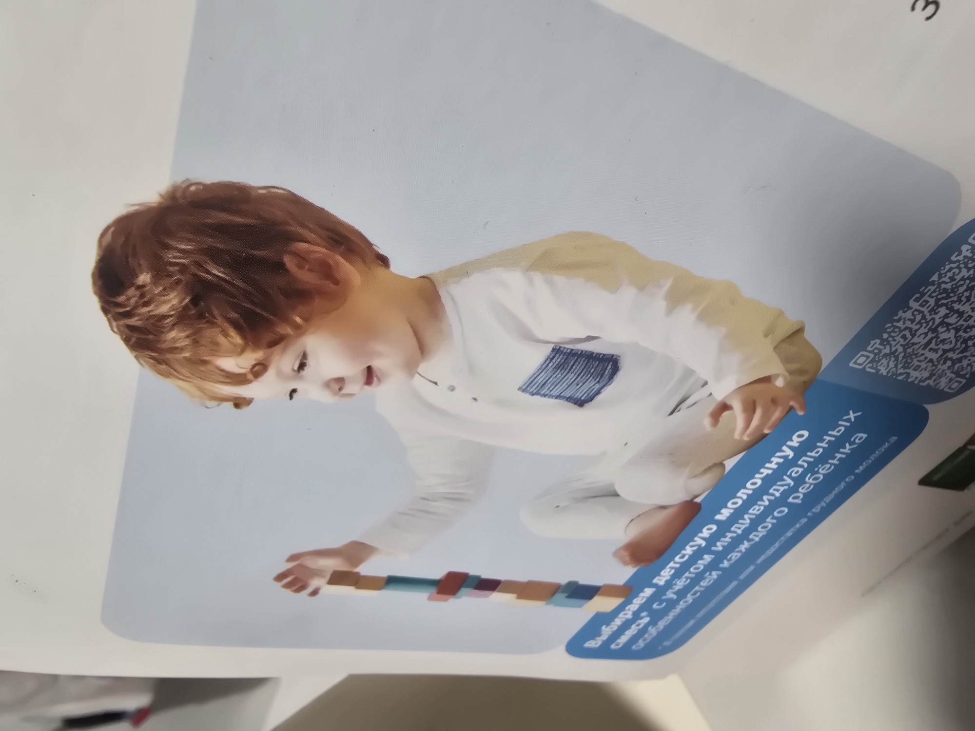 The image shows a child playing with wooden blocks. The child is a young boy with red hair, wearing a white shirt with a blue pocket and white pants. He is smiling and focused on building a tower with the blocks. The background is a light blue gradient. There is text in Russian on a blue banner at the bottom of the image, which translates to "Choosing milk formula * with individual characteristics of each baby." A QR code is also visible on the bottom right. The image is a photograph of a product advertisement or brochure. There is no information within the image to indicate a specific location in Ulan-Ude, Russia.