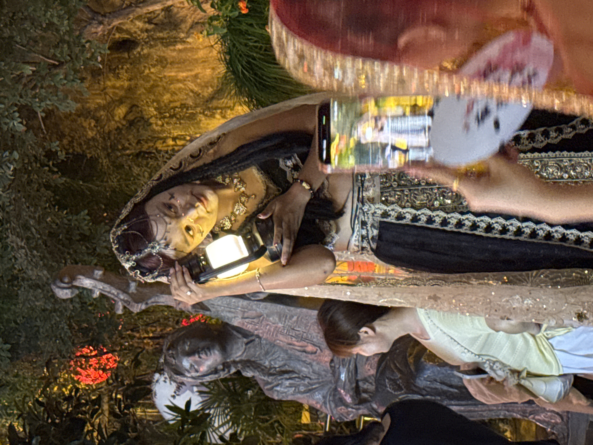 In Yuzhong District, China, at an outdoor nighttime event, a person dressed in ornate traditional attire, featuring a jeweled headdress and intricate gold and black garments, holds a glowing lantern. The individual's face is adorned with decorative markings. To the left, a stone sculpture of a person playing a stringed instrument stands amidst lush foliage and red illuminated decorations. In the foreground, another individual, facing away from the camera, is partially visible, holding up a smartphone to capture the scene, with a white fan held in their other hand. Further to the left, another person wearing a light yellow top and white pants is looking towards the sculpture. The overall atmosphere suggests a cultural performance or festival.