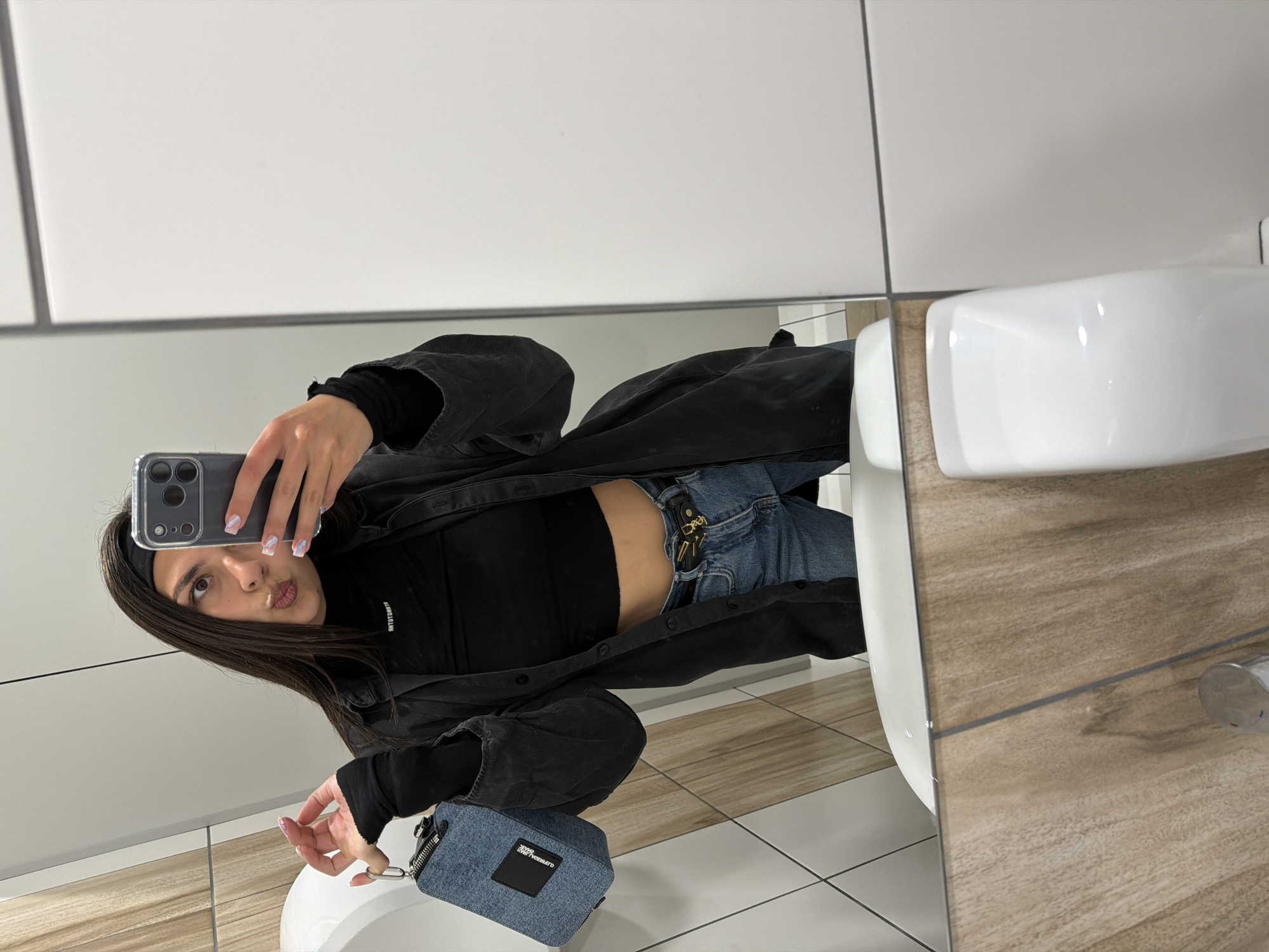 A woman is taking a selfie in a bathroom mirror. She is wearing a black turtleneck, jeans, and a dark overshirt or jacket. She has dark hair and is wearing a black headband. Her nails are painted with a marble pattern in white and light purple. In her left hand, she is holding a denim-like purse. The background shows a modern bathroom with white tiles and a wooden-effect countertop. There is a white sink in the foreground. The specific location in the city of Sofia, Bulgaria is not discernible from the image.