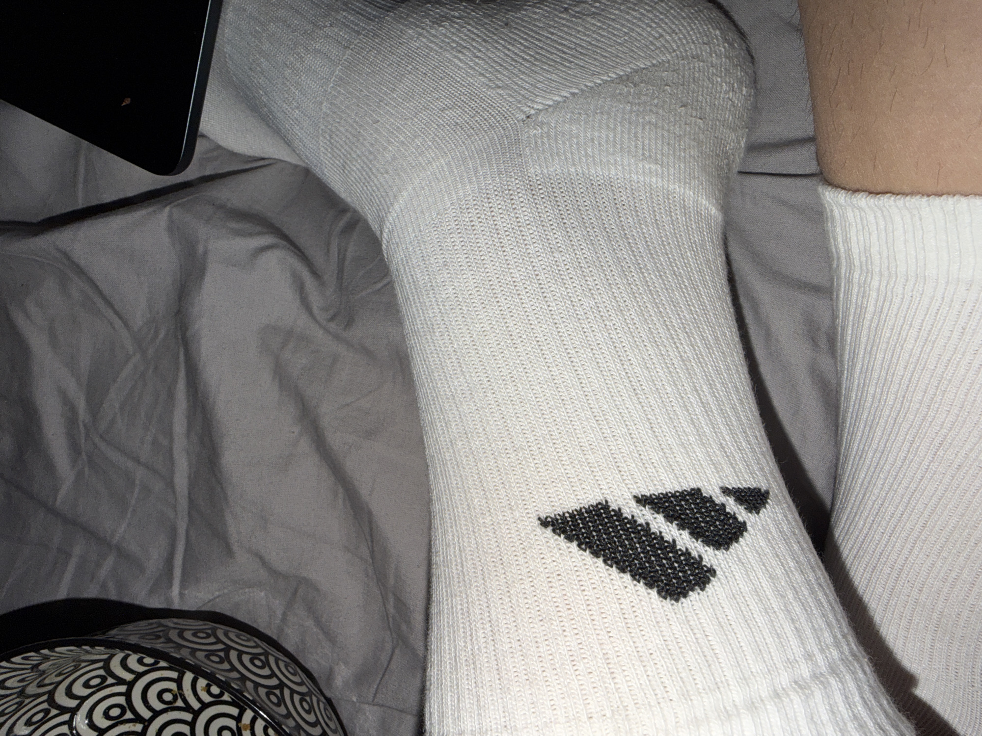 The image shows a close-up of a person's legs wearing white athletic socks with a black Adidas logo. The socks are on a bed with gray bedding. A patterned bowl is visible on the left side of the image, and a dark electronic device, possibly a tablet or laptop, is partially visible on the right. The scene appears to be indoors, in a domestic setting. There is no indication of the city of Oslo, Norway.