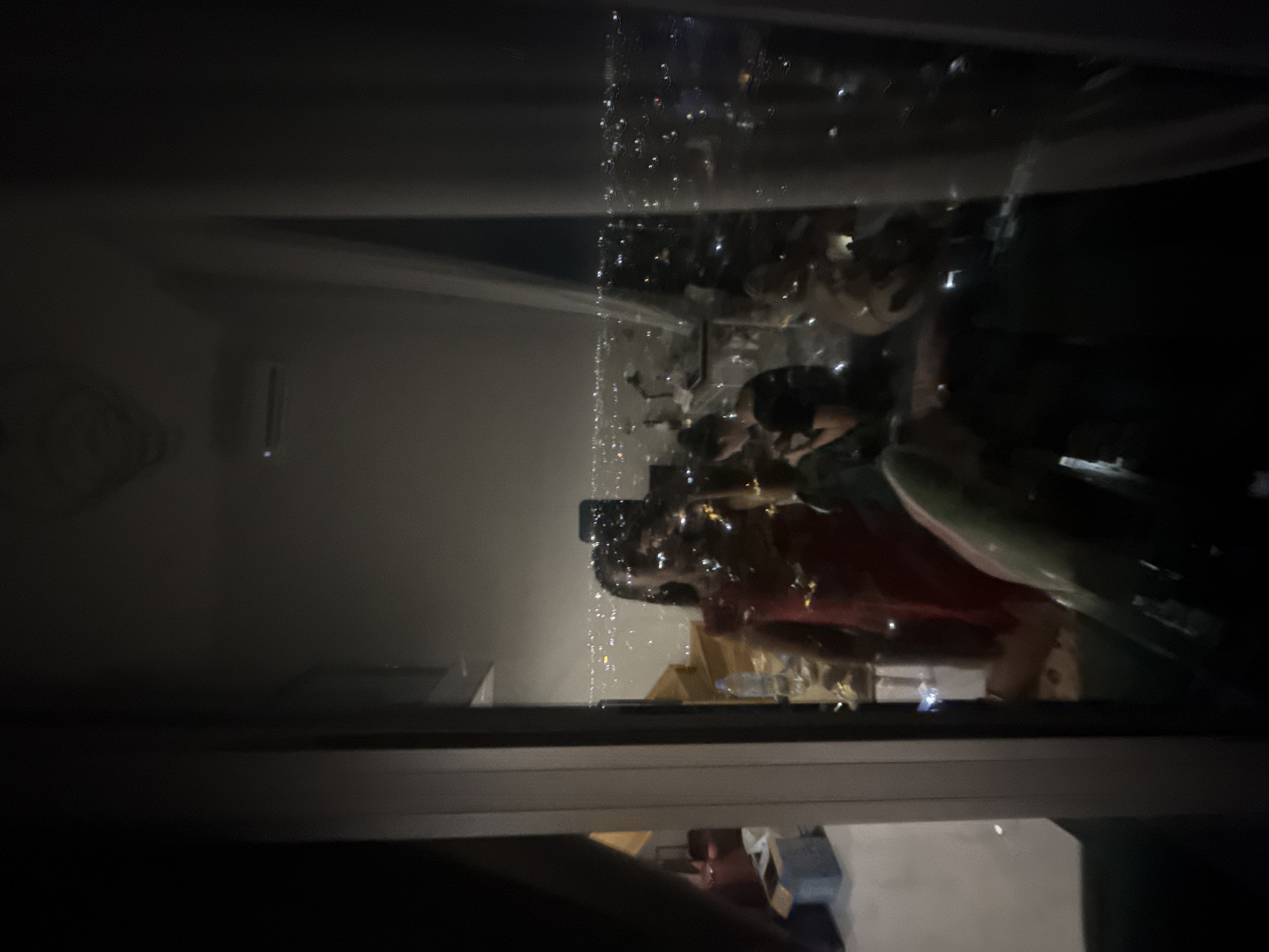 The image is a nighttime reflection in a large window, likely from a high-rise building. The foreground shows a partial reflection of a person wearing a red dress, holding a phone, and taking a selfie. To the right of this reflection, another person in darker clothing is visible. The background of the reflection is a blurred, illuminated cityscape, characteristic of a densely populated urban area, consistent with the location of Khan Mean Chey, Cambodia. Inside the room, some furniture and objects are dimly visible, including a staircase and a light fixture. The overall lighting suggests it is dark outside, with city lights being the primary illumination source for the reflected scene.