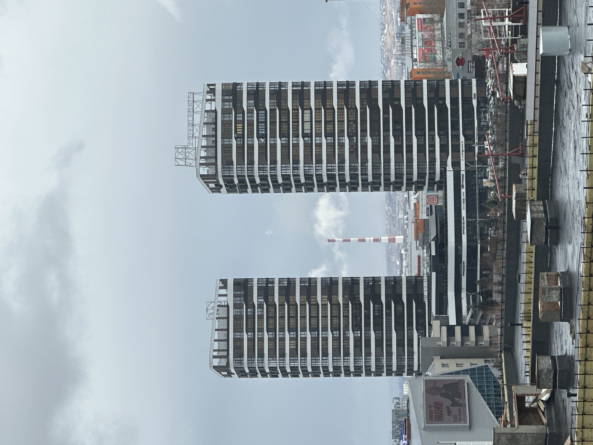 The image depicts two modern, high-rise buildings with a distinctive white and dark stripe design. The buildings are situated in an urban area, presumably in Krasnoyarsk, Russia, as evidenced by the "VIVALDI" advertisement. A red and white striped chimney can be seen in the background, suggesting an industrial presence in the vicinity. The foreground shows a construction site with construction materials, implying ongoing development in the area. The sky is overcast, and the overall atmosphere is somewhat industrial.