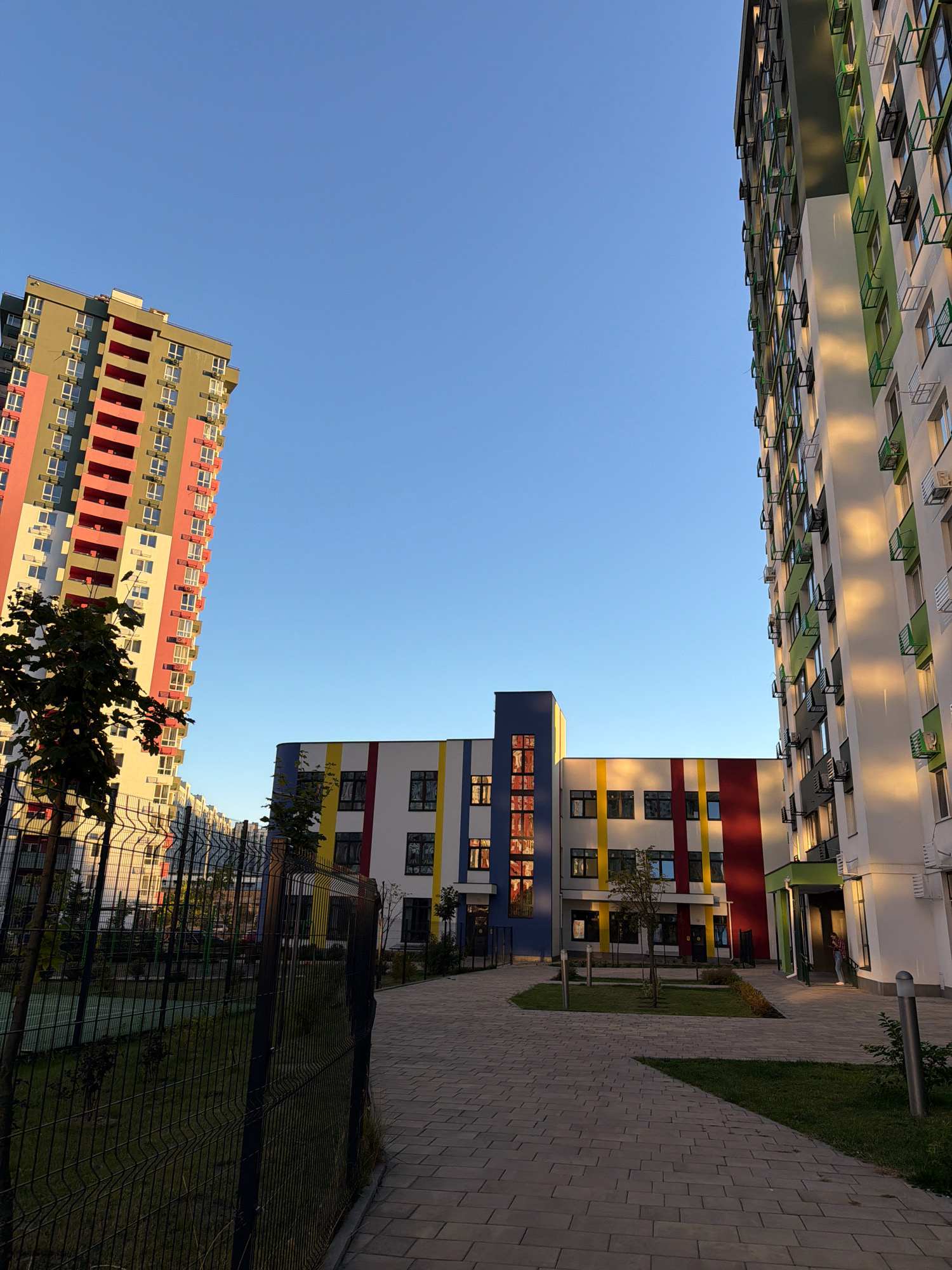This is a photograph taken in Kyiv, Ukraine, depicting a modern residential complex under a clear blue sky during what appears to be late afternoon or early evening, indicated by the warm sunlight casting long shadows and highlighting the buildings.
In the foreground, a dark metal chain-link fence runs diagonally across the frame, separating a grassy area from a paved walkway. The walkway is constructed of light gray interlocking pavers. Beyond the fence, a green lawn is visible, with a few young trees and bushes sparsely planted. A tennis court or sports area can be seen in the background through the fence.
Dominating the mid-ground is a low-rise building with a strikingly colorful facade. It features horizontal bands of white, yellow, red, and a dark blue section on the left. The building has numerous windows, some with dark frames.
To the left of the colorful mid-rise building, a tall, slender apartment building is angled towards the viewer. Its exterior is a mix of gray, pink, and white sections, with balconies featuring pink railings. Rows of windows line the facade.
On the right side of the frame, another tall apartment building stands prominently. Its exterior is predominantly white with sections of bright green and darker green accents. The building's facade is characterized by repeating windows and external air conditioning units. The setting sun casts a warm, golden light on this building, creating distinct vertical stripes of light and shadow.
A person is visible standing in the entrance of the tall white and green building on the right, partially silhouetted against the light. They appear to be wearing casual clothing.
The sky is a deep, clear blue, devoid of clouds, suggesting fair weather. The overall impression is one of a newly developed urban area with contemporary architecture.