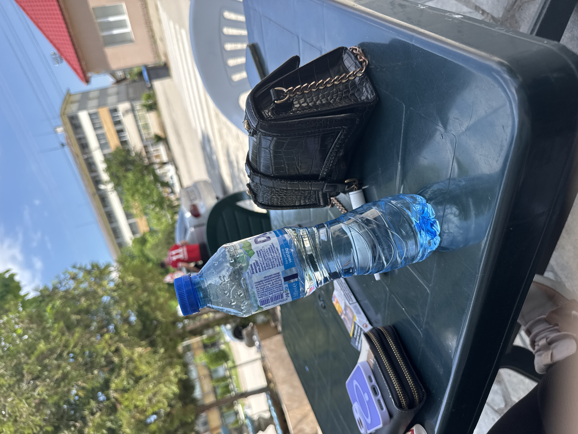 A slightly angled close-up shot captures a scene outdoors, likely in Drenovets, Bulgaria. Dominating the foreground is a dark green, possibly plastic, table. On this table rest several items. A partially visible black handbag with a crocodile skin pattern and chain details sits towards the top. A partially consumed clear plastic bottle of "Devin" brand natural mineral water lies near the center of the table. A light purple phone case is partly visible along with a wallet and possibly cigarette packs, indicating personal belongings.
The background features buildings and greenery typical of a city environment. A structure with a red roof is visible in the upper left, while taller buildings are seen further back, hinting at an urban setting. The image appears to be taken during daylight.
