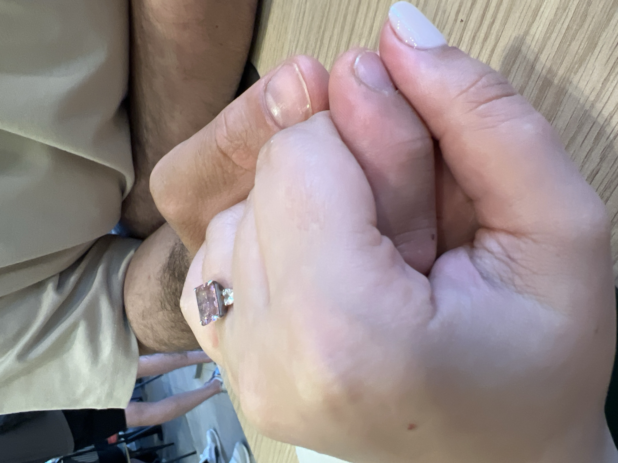 The image shows a close-up view of two people's hands clasped together. One hand, belonging to a person wearing a ring with a large, rectangular pink stone, is light-skinned with manicured nails. The other hand, belonging to a person with a visible patch of hair on their arm, appears to be darker-skinned or tanned. They are both wearing casual clothing, a light-colored t-shirt is visible. The background suggests an indoor setting with a wooden surface that could be a table. There are blurred figures of other people and shoes in the background, indicating a public space like a cafe or restaurant. There is no information or visual cue in the image that suggests a location in Yekaterinburg, Russia.