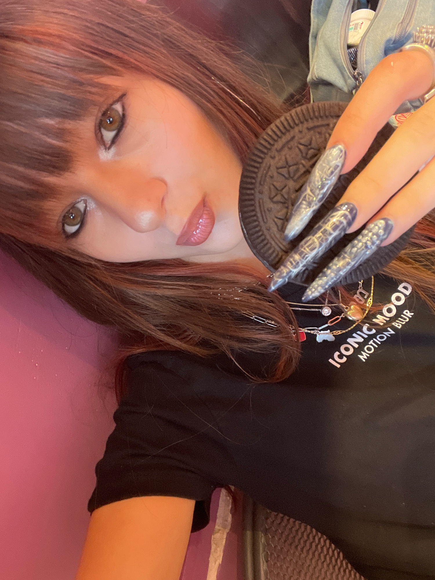 A person with brown hair and bangs is lying down and holding an Oreo cookie. They are wearing a black t-shirt with "ICONIC MOOD MOTION BLUR" written on it, and several necklaces. The person's nails are long and decorated with a silver, textured design. The background is a pink wall and a textured surface. There is no indication of the location being in Raanana, Israel.