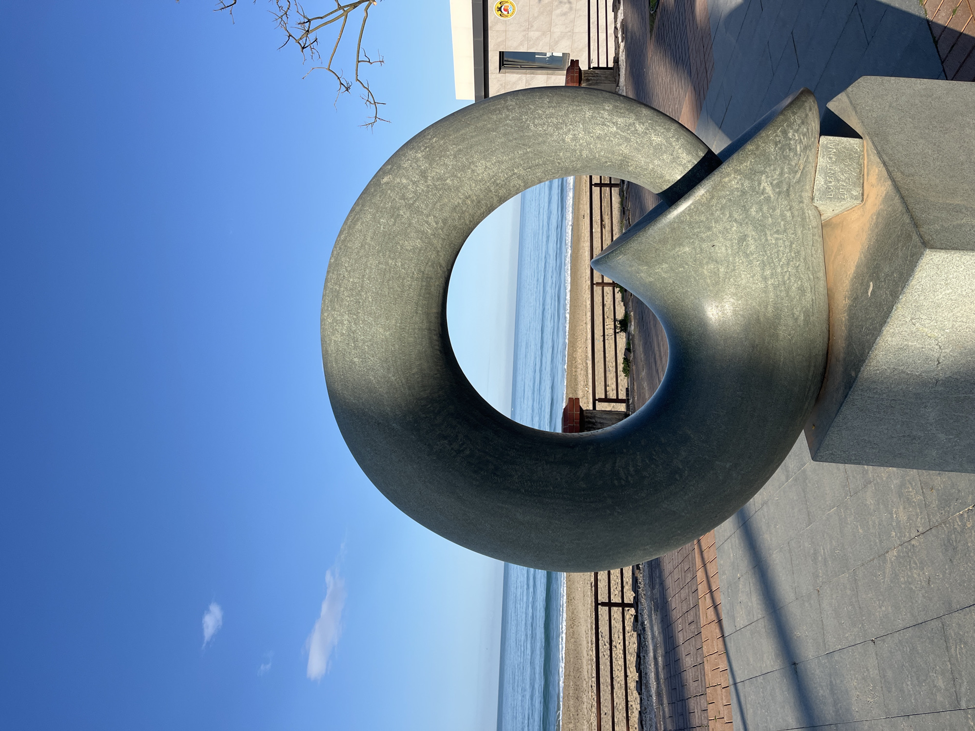 The image shows a large, gray, circular sculpture with an arrow-shaped extension, likely made of stone or concrete, standing on a brick and concrete paved area. The sculpture frames a view of the beach and ocean in Alanya, Turkey, with a blue sky above. The sculpture is located near a building and some bare tree branches are visible in the upper left corner. The picture is taken during the day with good lighting. The name "Bulent Ecevit" is visible on the statue.