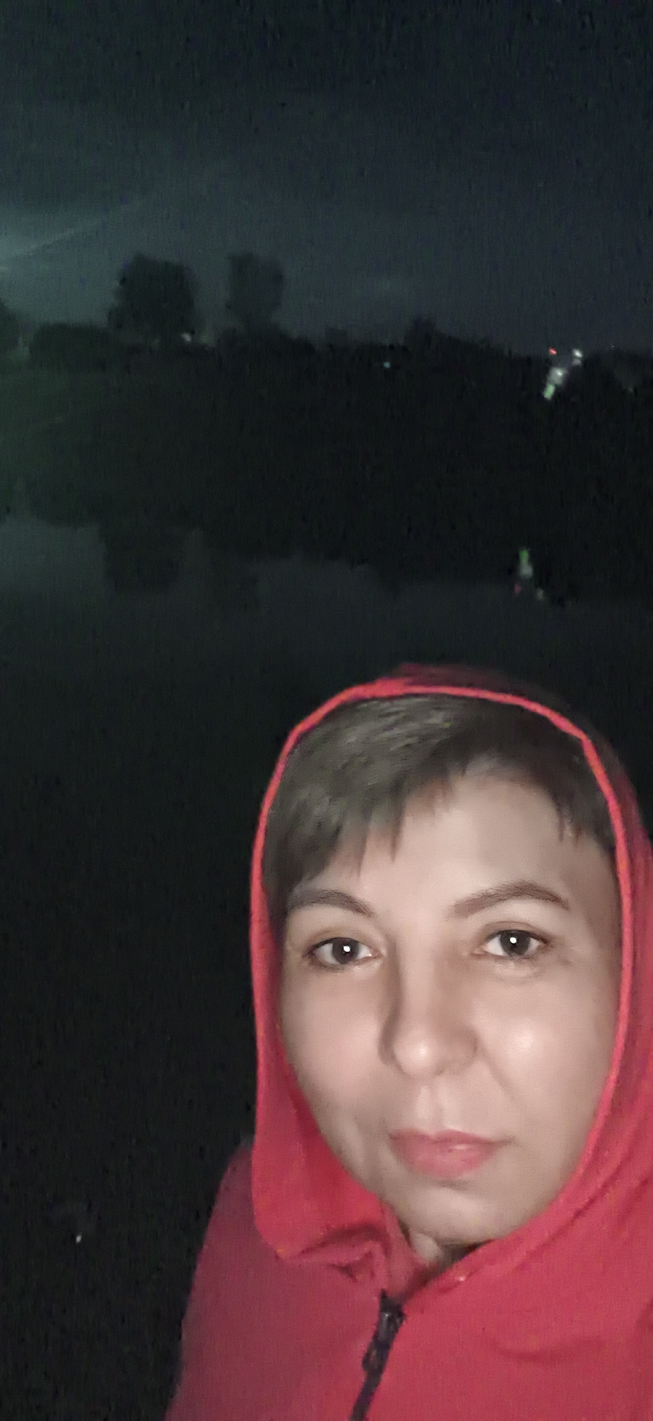 The image shows a close-up of a person's face, wearing a red hooded jacket. The background is dark and out of focus, with some blurred lights visible in the distance, suggesting an outdoor nighttime setting. The person's facial features are visible, including their eyes, nose, and mouth. The overall impression is of a selfie taken in low-light conditions. The location in the city of Фоки, Russia is not discernible from the image itself, as there are no identifying landmarks or context provided.