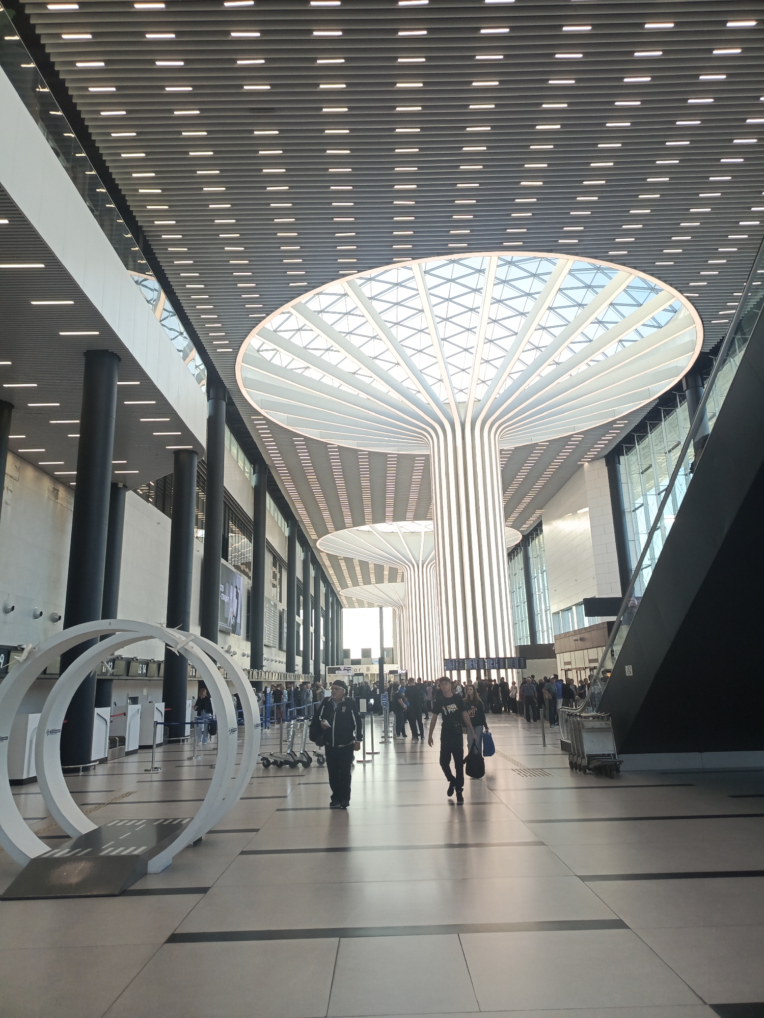 The image depicts the interior of a modern airport terminal. The architecture is characterized by a high, multi-level ceiling with a complex design featuring numerous linear lights and a large, open, circular skylight structure resembling a stylized tree or mushroom, with radiating beams of light. Supporting the ceiling are tall, dark cylindrical columns.
In the foreground, a man in dark clothing walks to the right, carrying a black bag over his shoulder. To his left, a white, circular sculpture with a ramp-like element is positioned on the polished floor. Further into the terminal, a queue of people is visible near check-in counters or gates. Several luggage carts are also present.
The environment appears clean and well-lit, with a sleek, contemporary aesthetic. The flooring is made of large, light-colored tiles with dark linear grout lines. Natural light enters from the skylight and large windows in the distance. The overall impression is one of a spacious and functional transportation hub. The location is identified as the city of Ob, Russia.