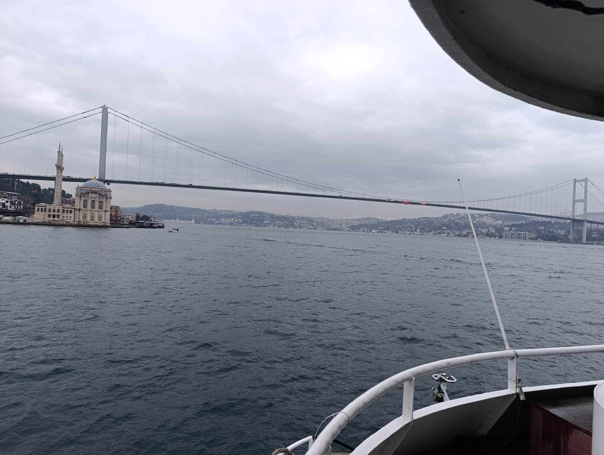 The image captures a view of the Bosphorus strait in Beşiktaş, Turkey. In the foreground, the railing and deck of a boat occupy the lower portion of the frame, suggesting the photograph was taken from the vessel. The water of the strait is visible, exhibiting dark, choppy waves. Across the water, the cityscape extends into the distance, partially obscured by a hazy or overcast sky. The iconic Bosphorus Bridge spans the strait, connecting the European and Asian sides of Istanbul. At the base of the bridge, on the European side, stands the ornate Dolmabahçe Mosque, a historical landmark. The overall atmosphere is muted due to the overcast weather conditions.