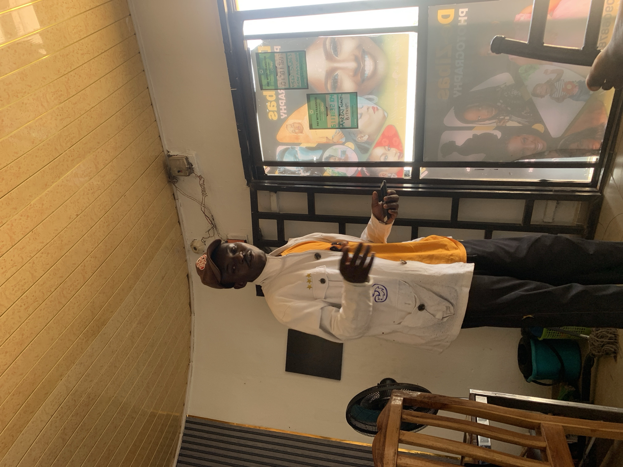 An indoor scene, likely a photography studio or shop, features a young, dark-skinned man standing centrally. He wears a brown baseball cap, a white long-sleeved jacket over a yellow shirt, and dark trousers. He holds a black smartphone in his raised right hand and looks towards the camera with a neutral expression.
To the man's left, a wall is covered with yellow and beige patterned paneling. To his right, a large window or glass partition displays promotional posters for a photography business. These posters feature smiling faces, primarily women and children, and include visible text. An old electrical outlet with exposed wiring is mounted on the wall near the window. Below the window, towards the bottom right, parts of a wooden chair, a black electric fan, a blue bucket, and a mop are visible. A dark rectangular object is affixed to the wall behind the man.
Visible text from the posters includes "PHOTOGRAPHY," "PAY WITH TRANSFER," and "OPay." Several numerical sequences, likely phone numbers, are present: "70017697," "056235963," and "6113832104." Names like "Bitkop Agron Nam" and "AARON NANYIL BITKOP" are also discernible, associated with payment or business information. The setting suggests a commercial space in Jos, Nigeria.