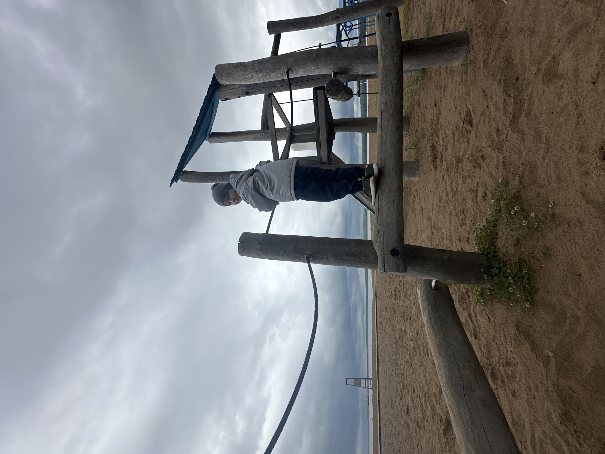 The image captures an outdoor scene at what appears to be a sandy beach or lakeside area in Chaikovsky, Russia, under an overcast sky.
In the foreground and midground, a young male child stands on a horizontal wooden beam of a rustic playground structure constructed from large, weathered wooden logs. The child wears a gray beanie, a light gray hooded jacket, dark blue trousers, and green and black athletic shoes. He is positioned facing slightly to his left, looking over his right shoulder towards the viewer with a slight expression. His hands are not clearly visible, possibly tucked into pockets.
The wooden playground structure features multiple vertical and horizontal logs, forming platforms and supports. A dark blue or green tarp-like material is draped over some logs at the top, suggesting a roof or partial shelter. A thick, dark rope is visible stretching horizontally from a wooden post on the left, curving downwards, and disappearing behind the child. A thinner black rope descends vertically from a higher platform on the right side of the structure. Partially obscured behind the child's left leg is a dark slide. The sandy ground around the base of the wooden posts features small patches of green vegetation with tiny white flowers.
In the background, a wide expanse of calm water, likely the Votkinsk Reservoir, is visible under the cloudy sky. A distant shoreline with a faint green line of land or trees marks the horizon across the water. On the sandy beach to the left, near the water's edge, a tall, A-frame wooden structure, possibly a lifeguard stand or observation tower, is present. A flat wooden walkway extends across the sand towards the water. Faint, dark shapes on the water in the far distance may indicate boats or other structures. On the far right edge of the frame, partially obscured blue structures suggest additional playground equipment or shelters. The sky is uniformly gray with diffuse cloud cover.