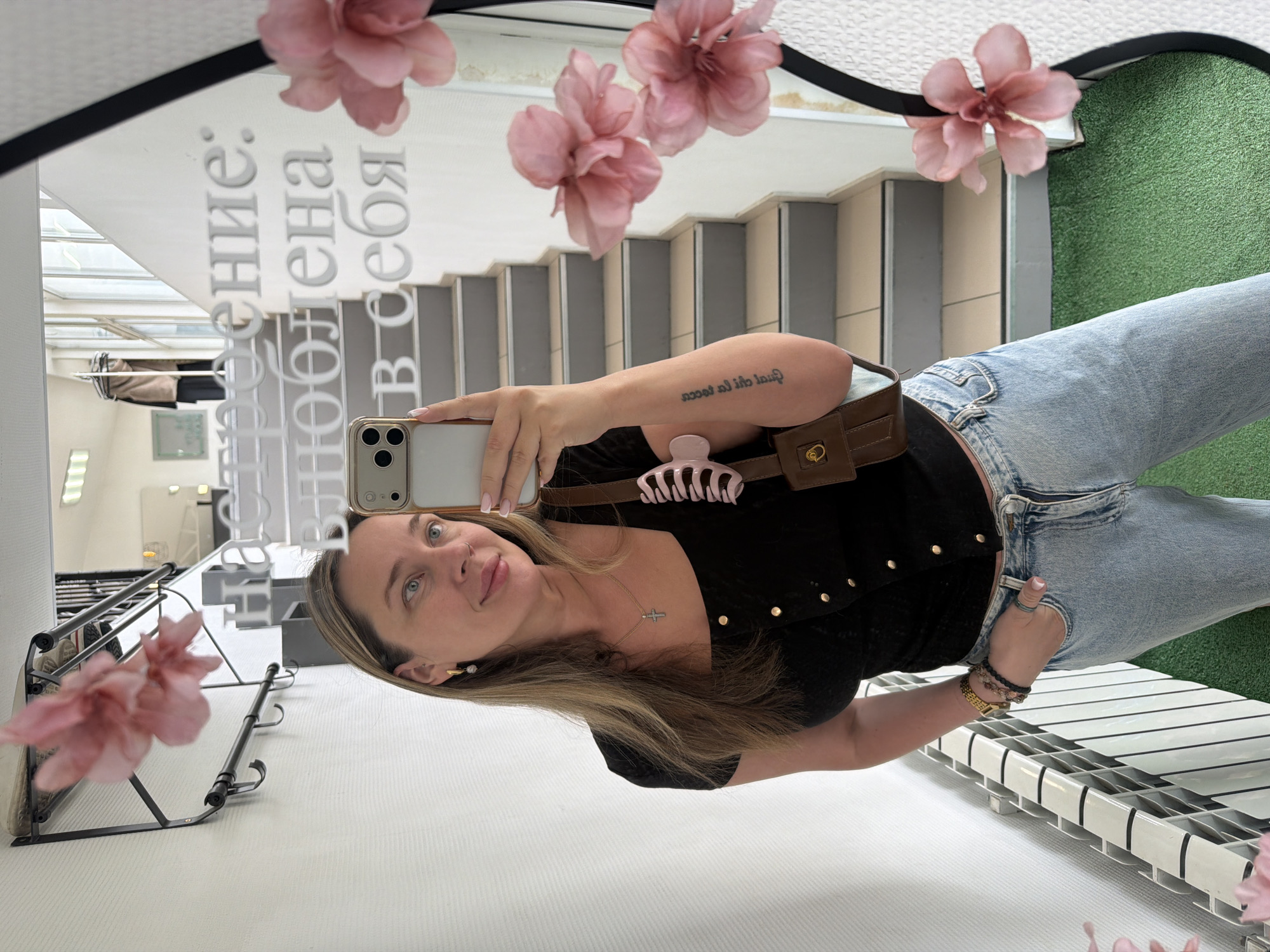 An indoor mirror selfie captures a woman with light brown hair and blue eyes, looking towards the camera with a subtle smile. She is wearing a black short-sleeved top with gold studs, light blue denim jeans, and a brown waist bag adorned with a pink hair claw clip. A silver cross necklace, a gold watch, and bracelets accessorize her outfit. She holds a light-colored smartphone to take the photo, and a tattoo reading "amour de ma vie" (French for "love of my life") is visible on her right forearm.
The background, reflected in the mirror, reveals a multi-level interior space. A staircase descends behind her, and a section of artificial green turf is visible on a wall. Several delicate pink artificial flowers are attached to the mirror's frame, with one prominently in the foreground. Other background elements include a white shelving unit and a black metal rack with hooks.
White Cyrillic text is prominently displayed on the mirror's surface, reading "ОТНОШЕНИЕ: ВЛЮБЛЕНА В СЕБЯ," which translates to "ATTITUDE: IN LOVE WITH MYSELF." The scene is brightly lit, suggesting daytime. The setting appears to be a commercial establishment or a stylish personal space in Chaikovsky, Russia.