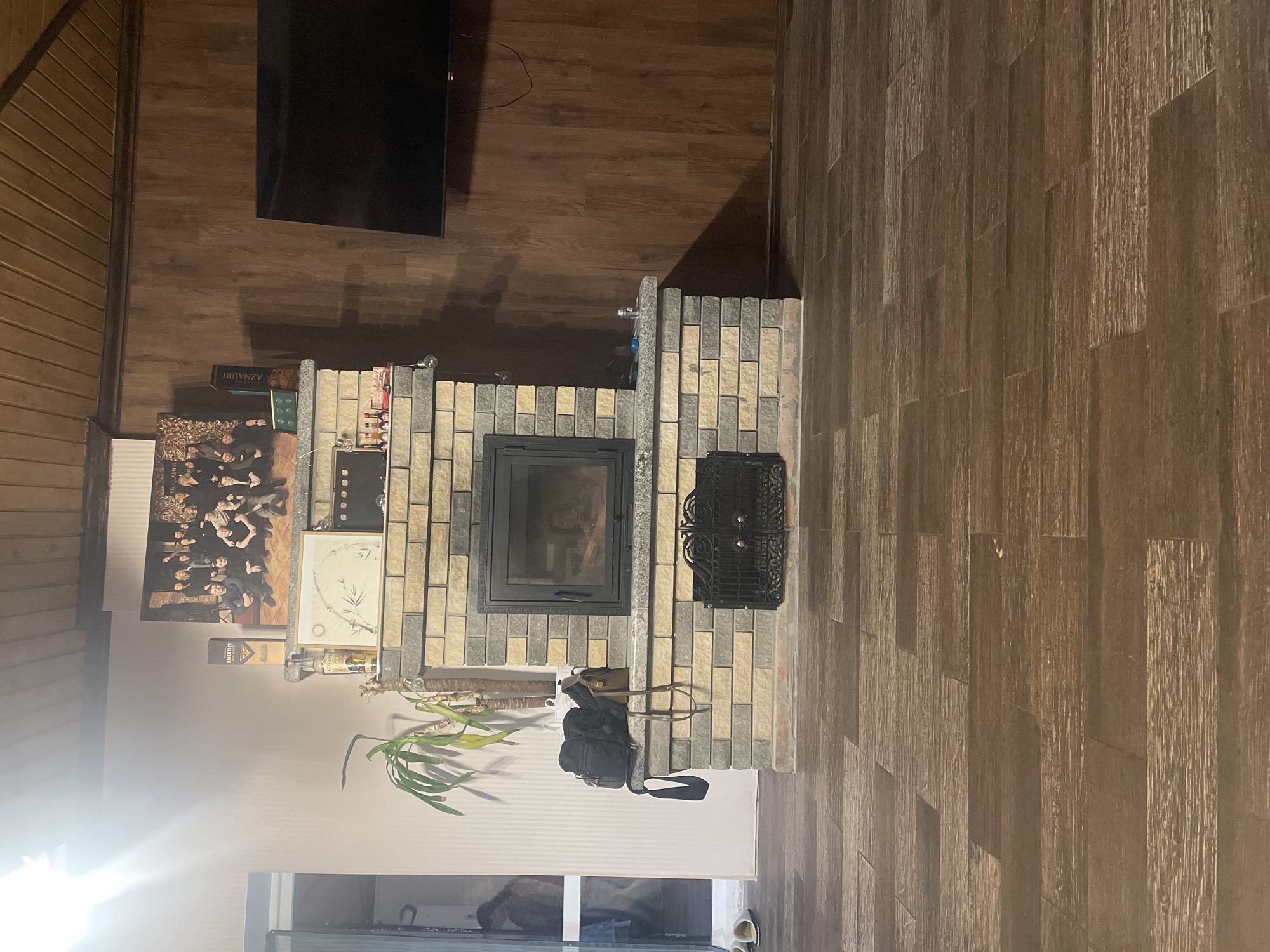 The image depicts an indoor domestic space, likely a living room, in Petrivka, Ukraine, characterized by a central brick fireplace, a mix of wall finishes, and dark wood flooring. **Environment:** The floor is covered with dark brown, wood-patterned tiles arranged in a staggered horizontal pattern, simulating distressed wood planks. The left wall is painted a light pinkish-beige with a subtle vertical striped texture, while the right wall and ceiling are clad in dark brown wooden paneling, with the ceiling featuring horizontal slats. A darker wooden beam runs along the junction of the ceiling and the right wall. An open doorway on the far left reveals an adjacent room; two white slippers are on the floor near its threshold. **Central Objects:** A prominent fireplace, constructed with alternating light beige and dark gray rectangular bricks, serves as the focal point. It features a light gray, granite-like mantle and hearth. A black metal, glass-fronted fireplace insert is embedded in the brickwork, displaying active flames or embers within. Below the main firebox, a decorative black metal grate covers an opening, likely an ash pit or storage area. **Objects on and Around the Fireplace:** Above the fireplace, on the light-colored wall, a large framed photograph depicts a group of approximately ten to twelve individuals, primarily men, dressed in dark attire. On the upper fireplace mantle (shelf), a tall green houseplant in a white pot is situated on the left, alongside a black backpack and a brown shoulder bag. On the right side of the mantle, a small clear glass jar, a blue object (possibly a lighter), and part of a string light are visible. The lower mantle (hearth) displays a framed picture with a circular bamboo motif. To its left are two bottles: one labeled "LIBERTIES The Gin" and another, partially visible, labeled "AZNAURI." To the right of the framed picture, a small gold frame contains four green circular objects, and a collection of miniature liquor bottles rests on a small tray. **Other Features:** On the right wood-paneled wall, a large, dark flat-screen television is mounted and is currently off; associated cables are visible beneath it. The room is illuminated by ambient light, with a strong glare emanating from an unseen light source in the upper left corner, possibly from a window or a bright overhead fixture. **People/Actions:** No individuals are distinctly visible in the immediate foreground. A blurred, indistinct figure or reflection of a