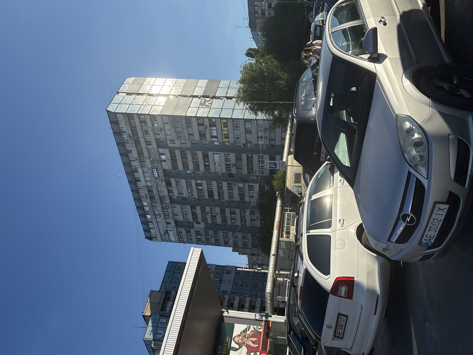A sunny day in Dnipro, Ukraine, captures a street scene with parked cars, a gas station, and apartment buildings. A silver Opel sedan is in the foreground, its license plate reading "AE 3002 IX". To its left, a white Skoda station wagon with license plate "AE 5087 OT" is parked. Behind these vehicles, a dark SUV is visible. A gas station canopy with a billboard featuring a woman is on the left side of the image. In the background, a tall, weathered apartment building dominates the skyline, its windows reflecting the sunlight. Several other apartment buildings, some with blue facades, are also present, along with trees and telephone poles. The sky is a clear blue.