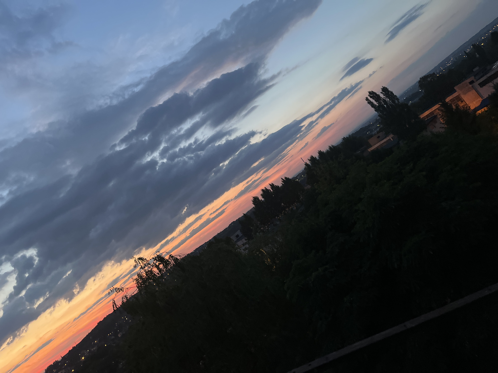The media file displays a landscape at dusk, with a prominent diagonal composition. The sky occupies the upper portion of the frame, showcasing a gradient of colors from deep blue at the top to warm orange and pink hues near the horizon, indicating sunset or sunrise. Dark, dramatic clouds are scattered across the sky, creating depth and texture.
Below the sky, a silhouetted treeline runs along the middle ground, with some trees appearing taller and more distinct than others. Beyond the trees, hints of a city or town are visible in the distance, with scattered lights suggesting it is nighttime or early evening. Some buildings with illuminated windows are also discernible.
The foreground consists of dark, dense foliage, obscuring the ground and any potential objects or people. The overall mood is serene and contemplative, evoked by the soft twilight colors and the quietude of the scene. The weather appears calm, with no strong indications of wind or precipitation. The image's diagonal tilt adds a dynamic element to the otherwise still landscape. No text is visible in the media file.