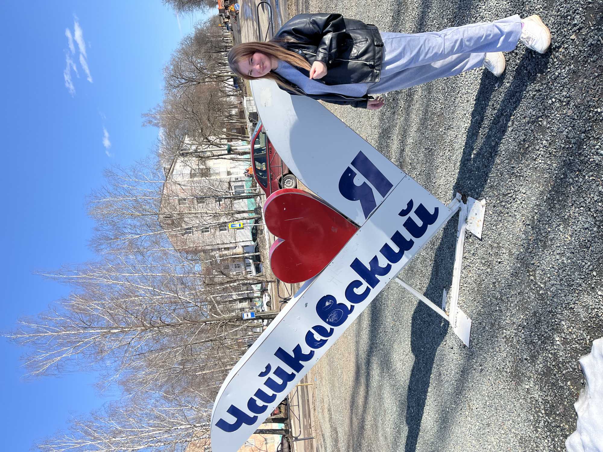 A young woman is leaning on an I-heart-Chaikovsky sign in the Russian city of Chaikovsky. The sign is white with blue Cyrillic text spelling "Chaikovsky" with a red heart. The woman is wearing a black leather jacket, grey sweatpants, and white sneakers. She has long brown hair and is smiling. The background includes bare trees, a light blue sky with wispy clouds, a multi-story building, and a parked car. The ground is covered in gravel.